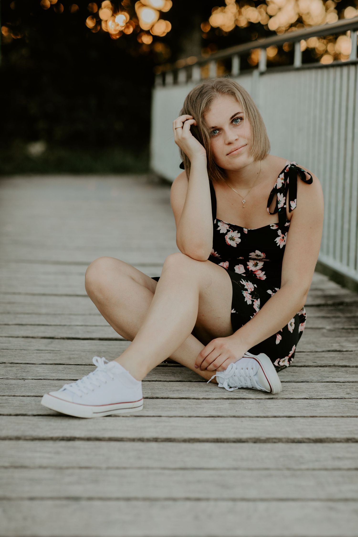 Person in floral dress sitting on wooden deck wearing white sneakers at golden hour.