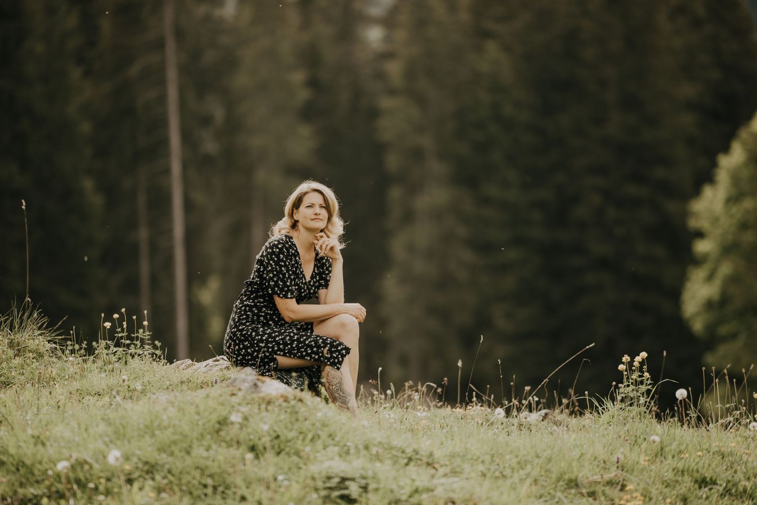 A person in a dark dress sits in a sunlit meadow with forest behind.