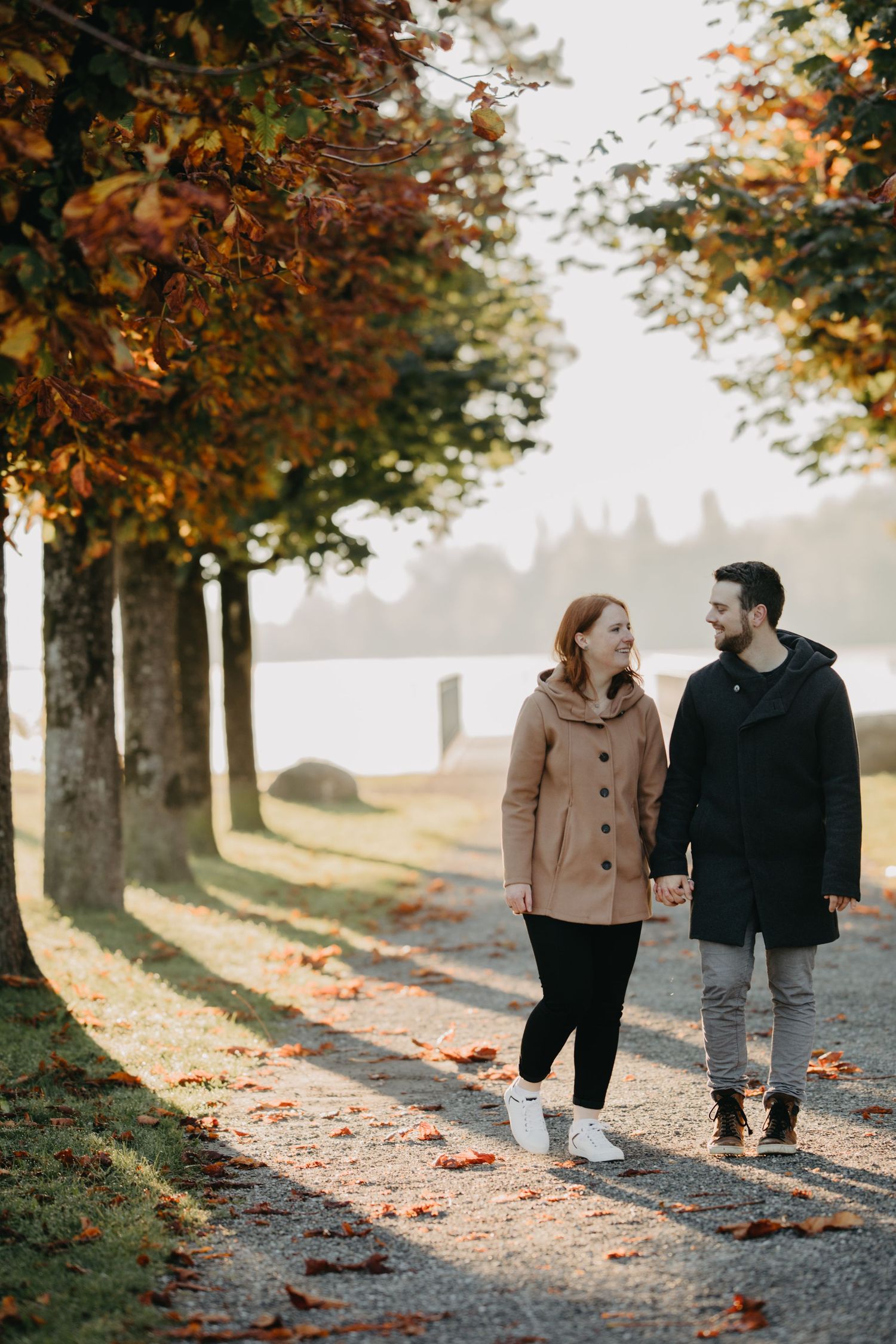 Autumn path lined with trees as couple walks beside lake.