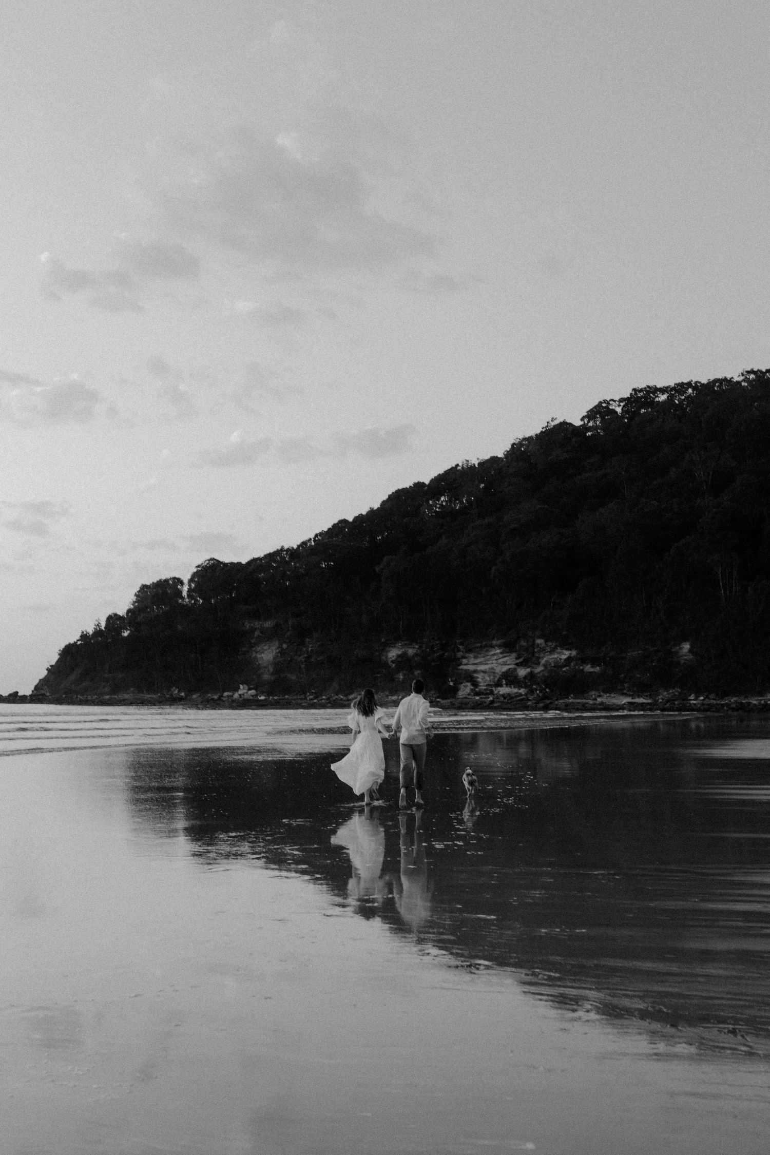 Silhouetted figures walking on reflective beach with mountains in background.