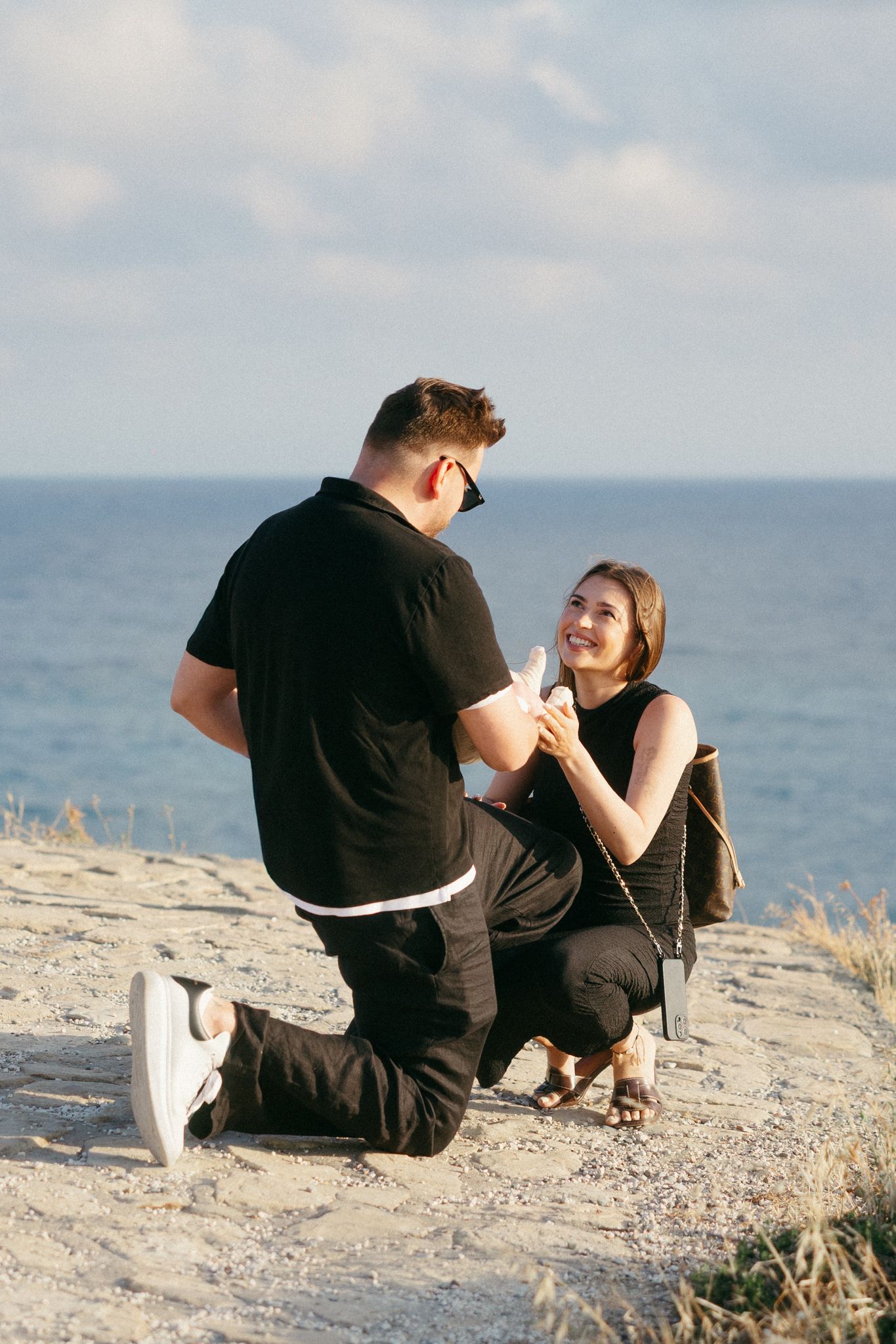 Man proposes to smiling woman on rocky seaside cliff, both dressed in black, with ocean backdrop.