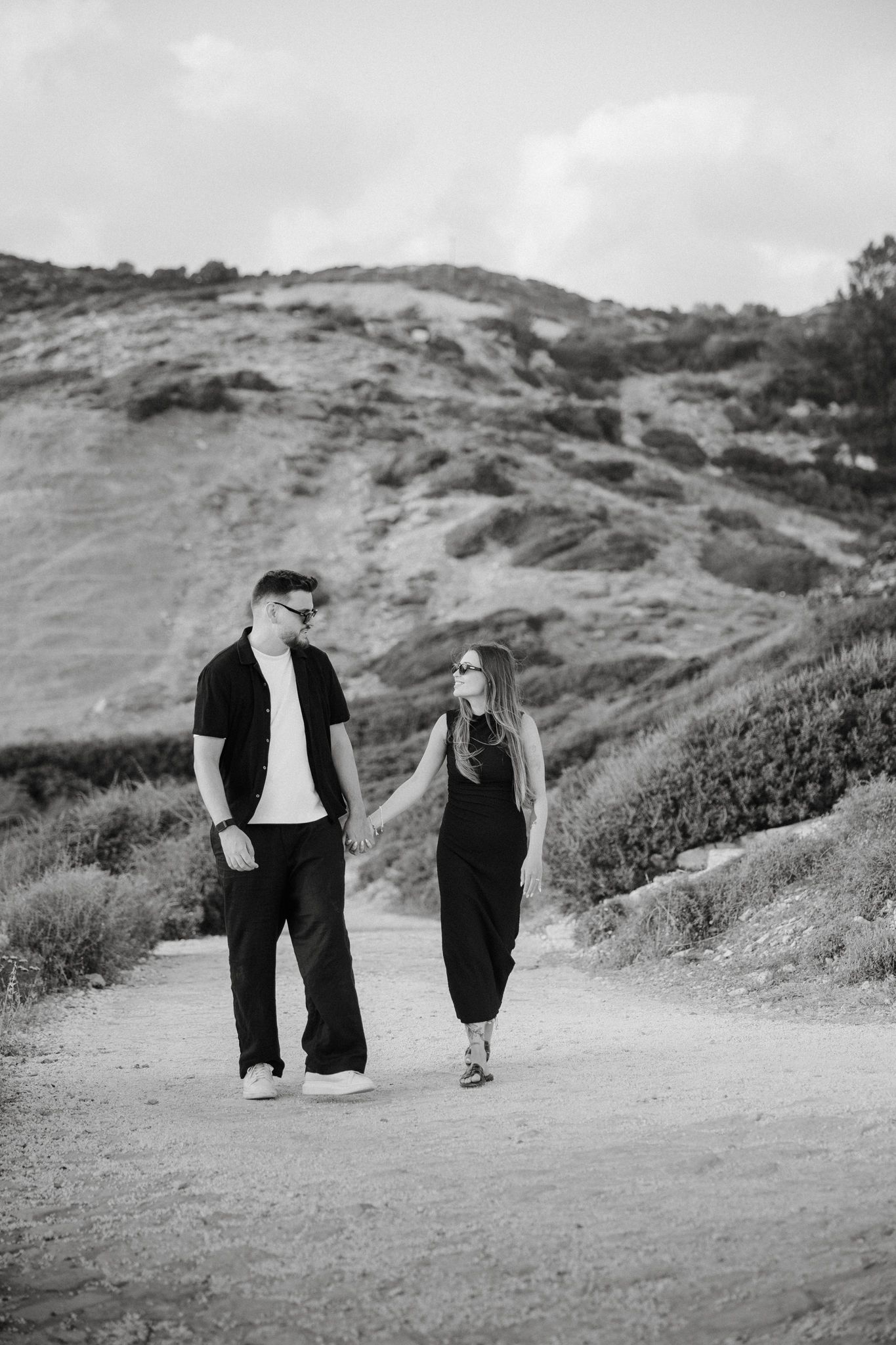 Black and white photo of a couple holding hands while walking on a dirt path with a scenic hillside backdrop.
