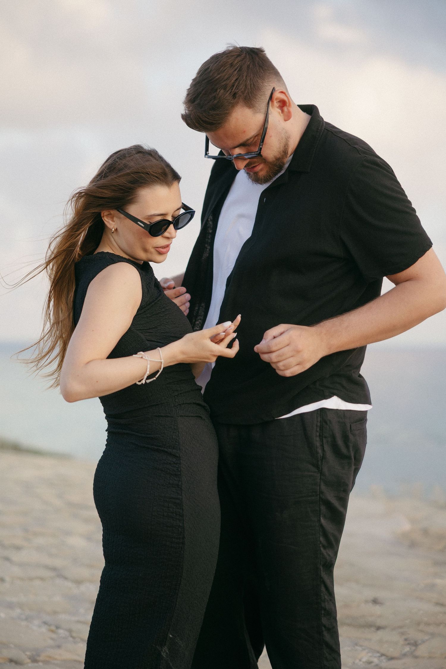 Couple in black outfits and sunglasses standing on a beach, woman touching man's shirt in a romantic pose.
