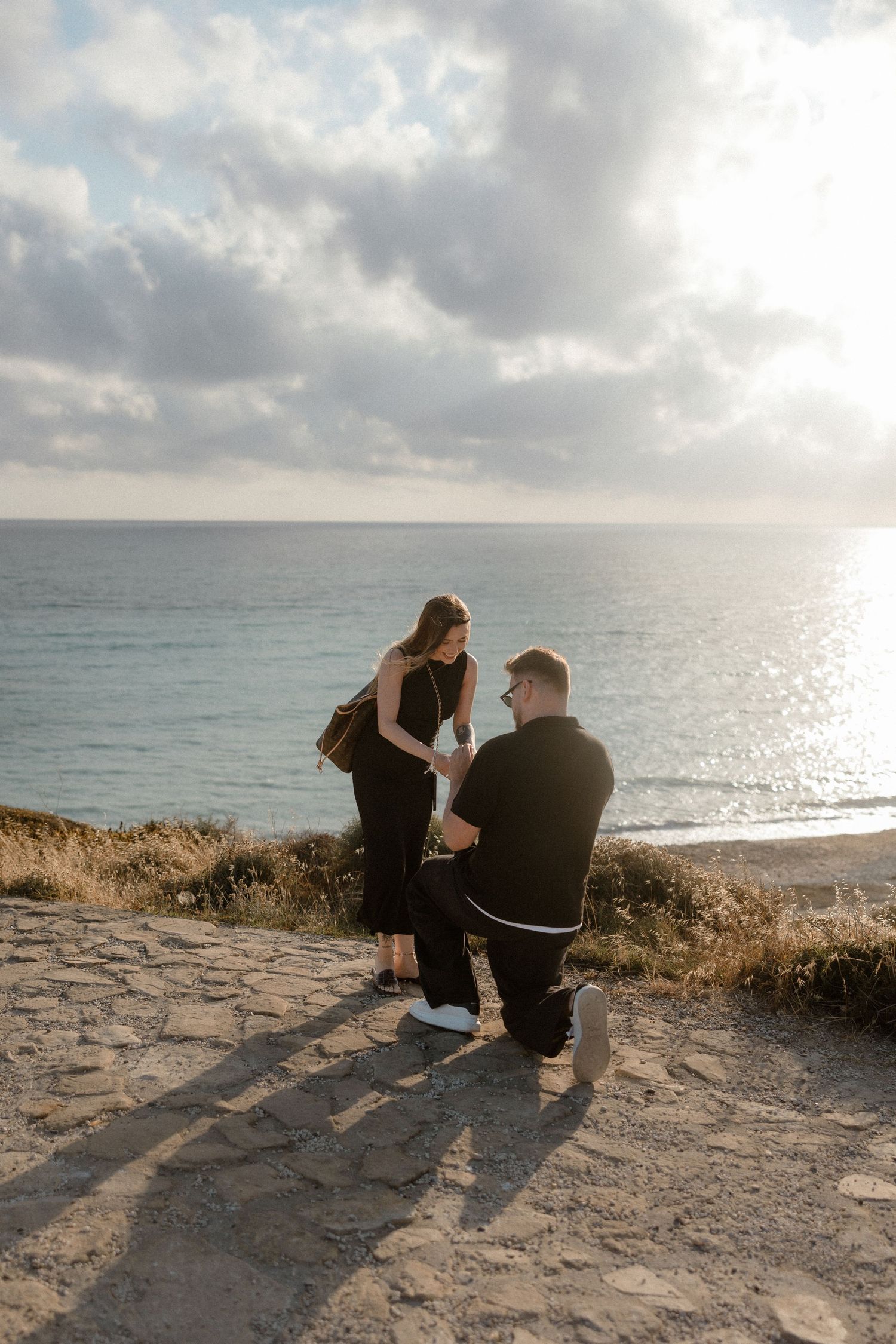 Man proposes on one knee to woman on scenic coastal cliff overlooking ocean at sunset.