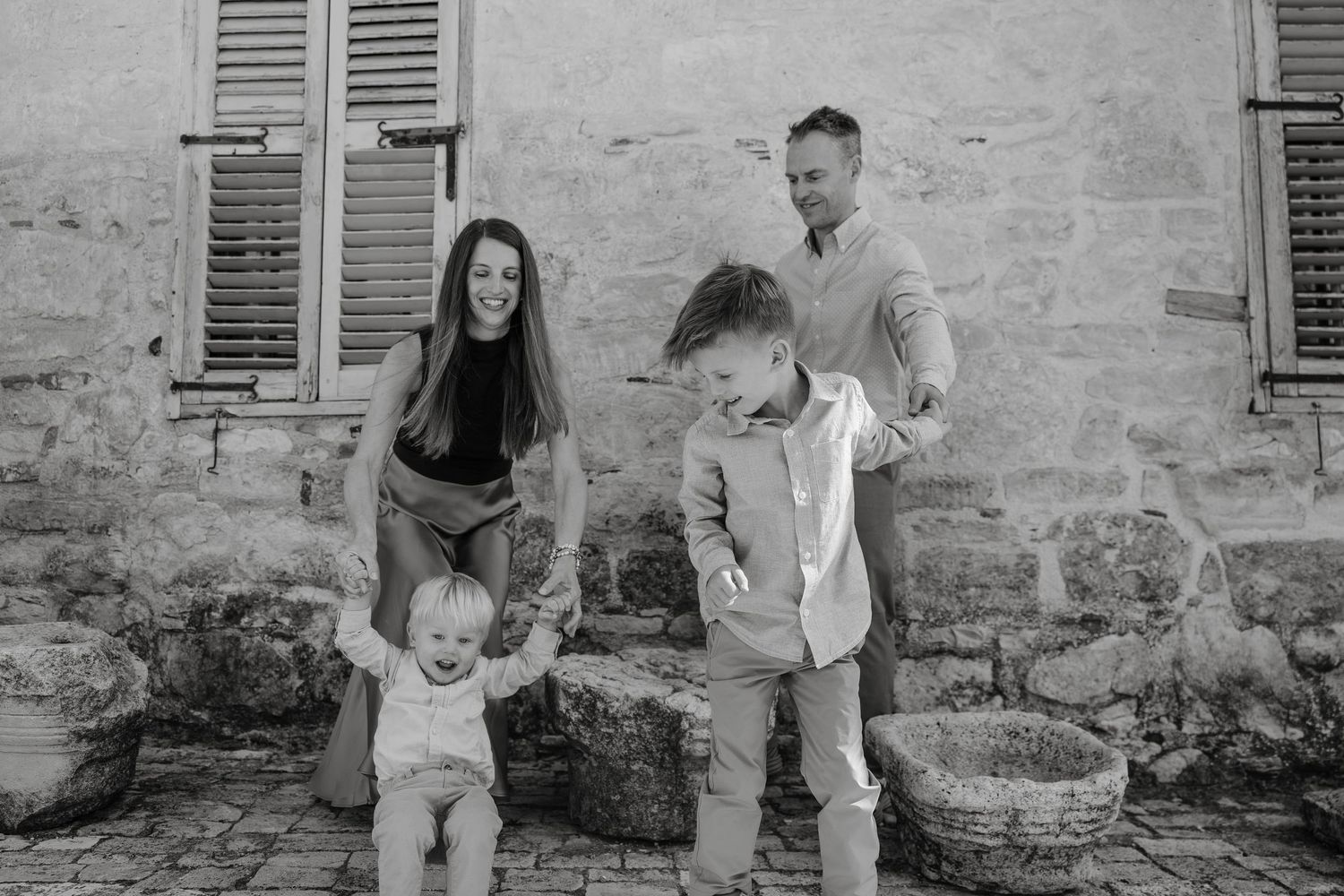 Black and white family photo of parents with two young children on stone steps near rustic wall with shuttered windows.