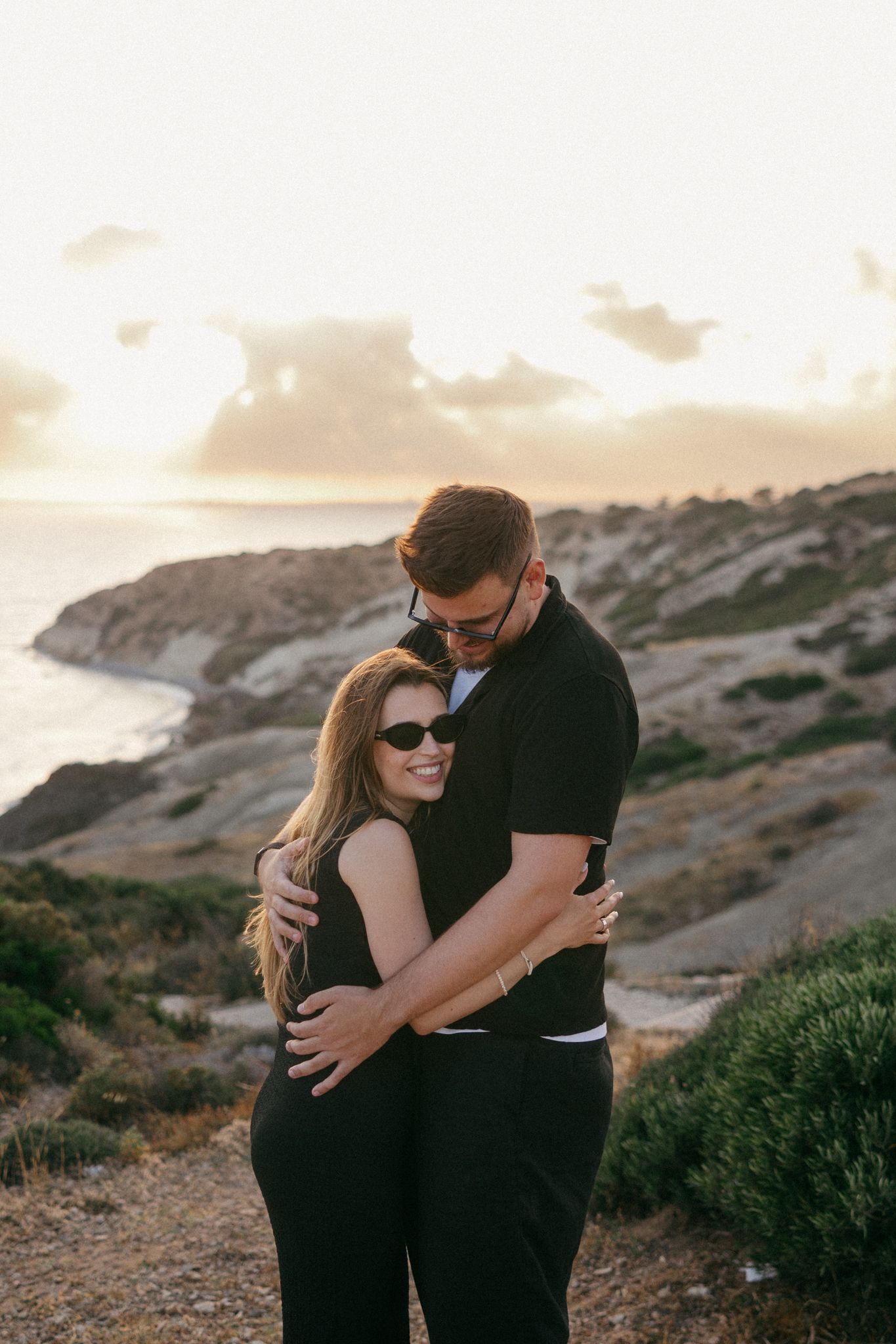 Couple embracing at sunset on rocky coastal cliffside, both wearing black outfits and sunglasses, ocean view background.