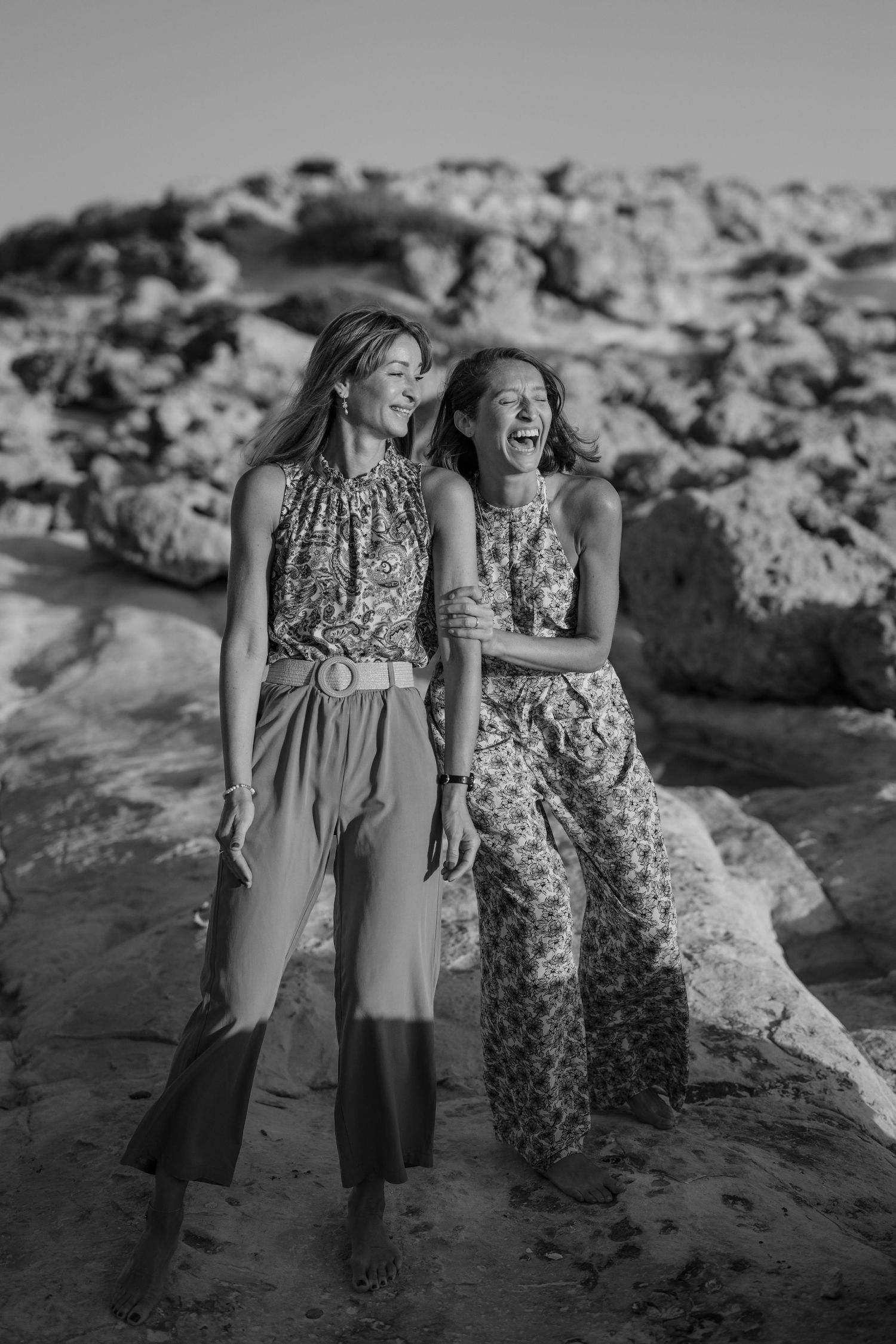 Two women laughing together on rocky beach in black and white photo, wearing floral and patterned outfits.