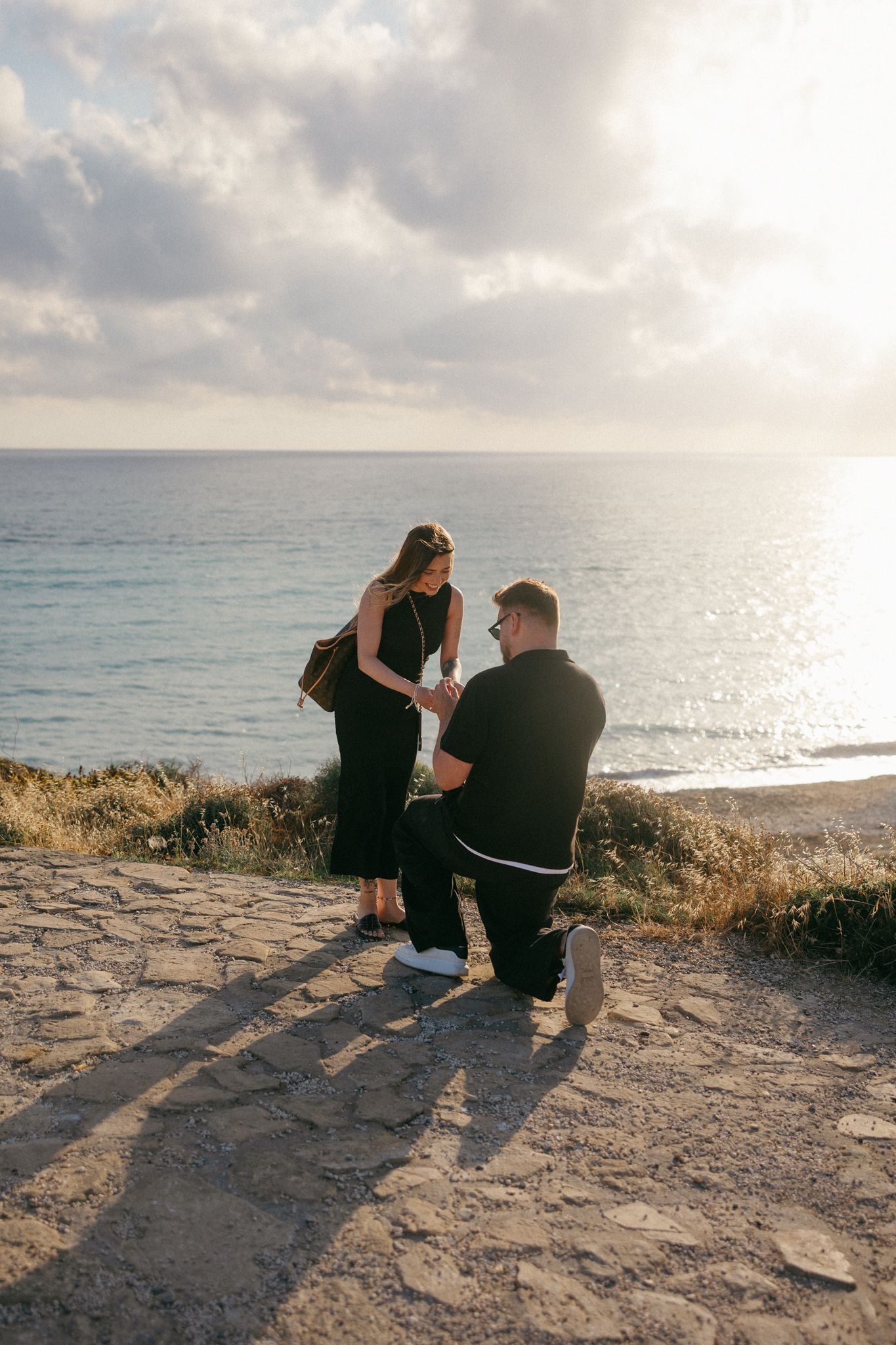 Man proposes on one knee to woman on scenic coastal cliff at sunset, both dressed in black.