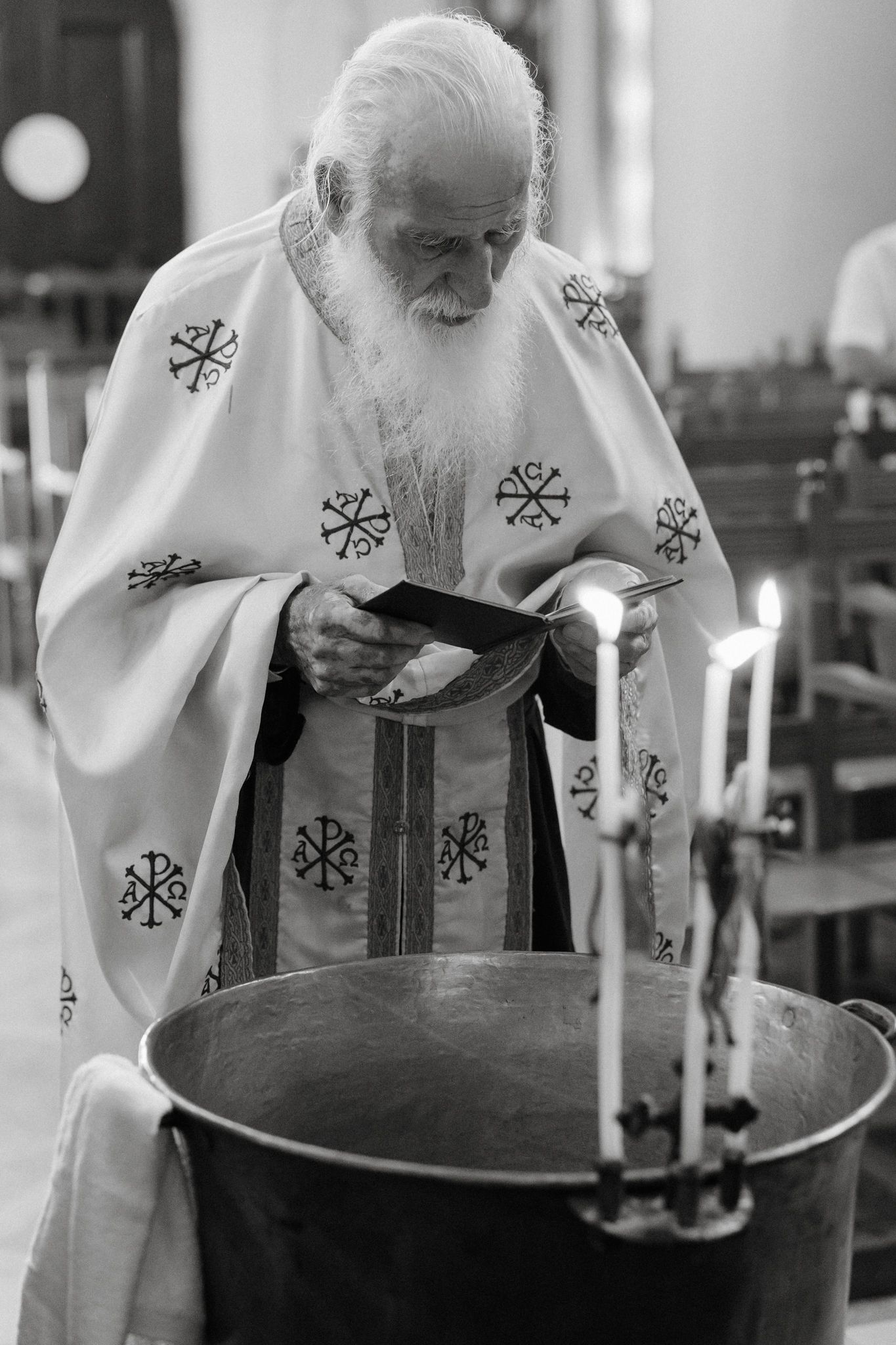 Elderly Orthodox priest in white vestments reads scripture over baptismal font with lit candles in black and white.