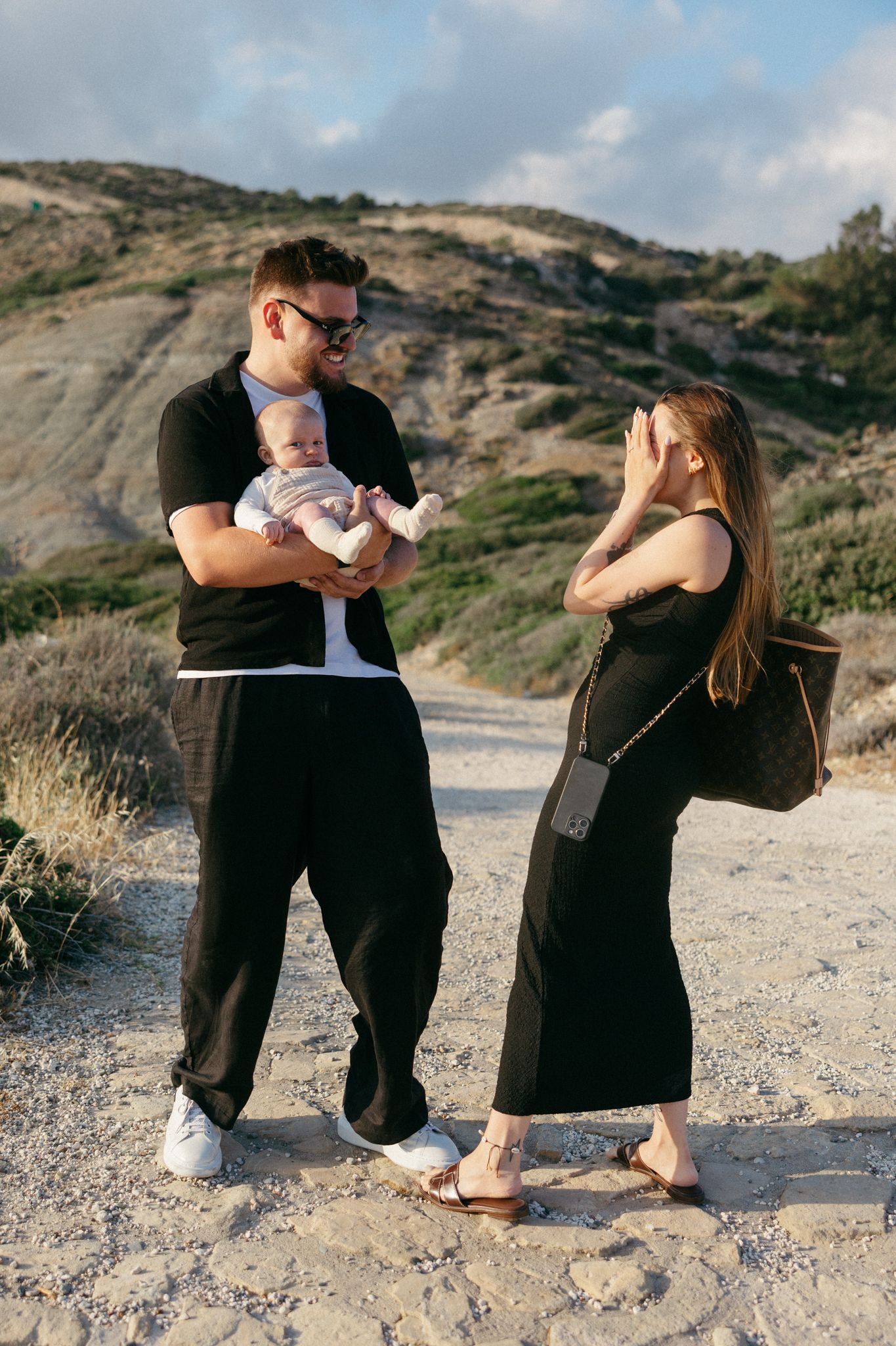 Young family with baby outdoors on rocky trail, man in black holding infant while woman shields eyes from sun.
