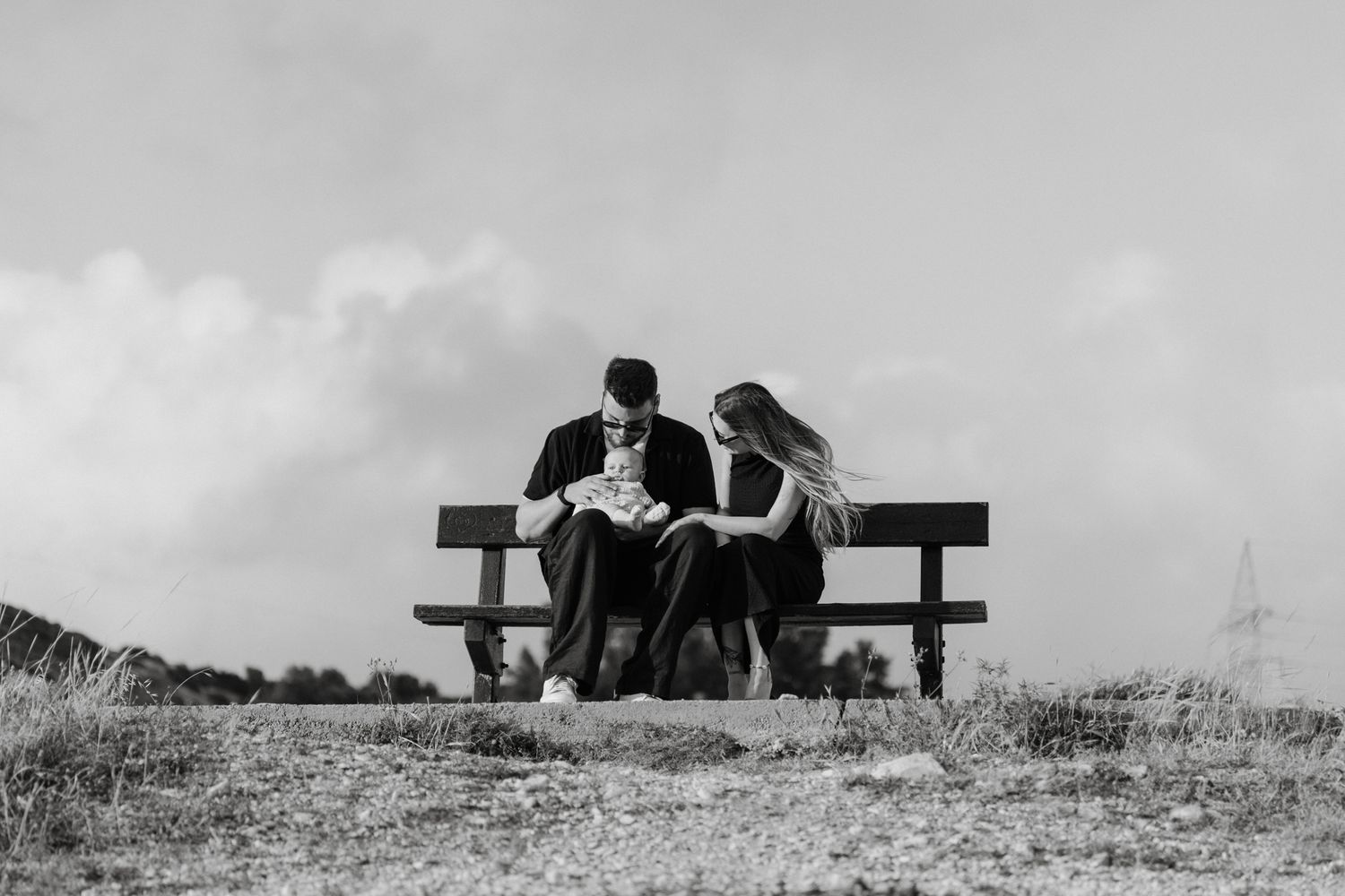 Couple sitting close together on a wooden bench outdoors, reading in a serene black and white landscape.