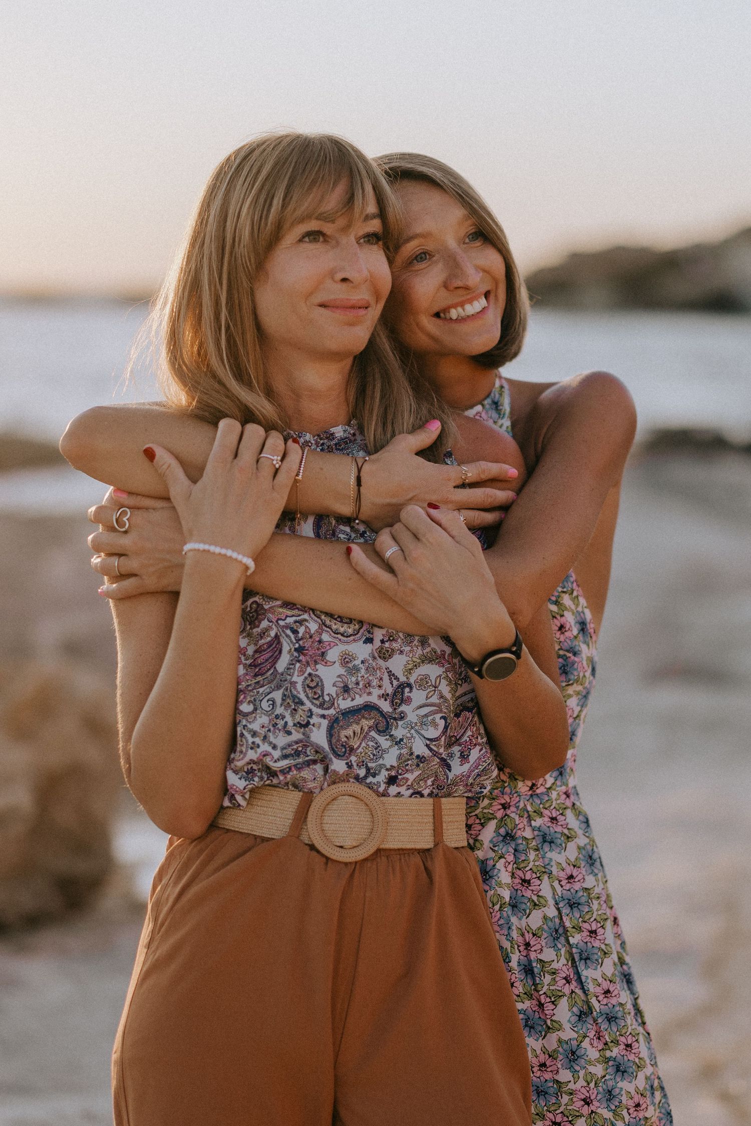 Two women hugging and smiling on a rocky beach at sunset, wearing floral and bohemian summer outfits.