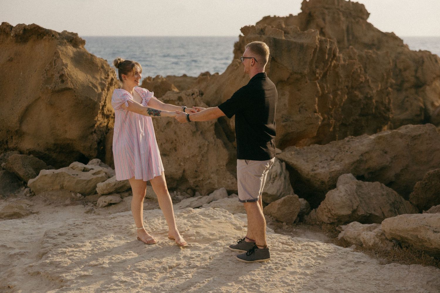Couple holding hands and dancing on a rocky beach at sunset, woman in pink dress, man in black shirt.