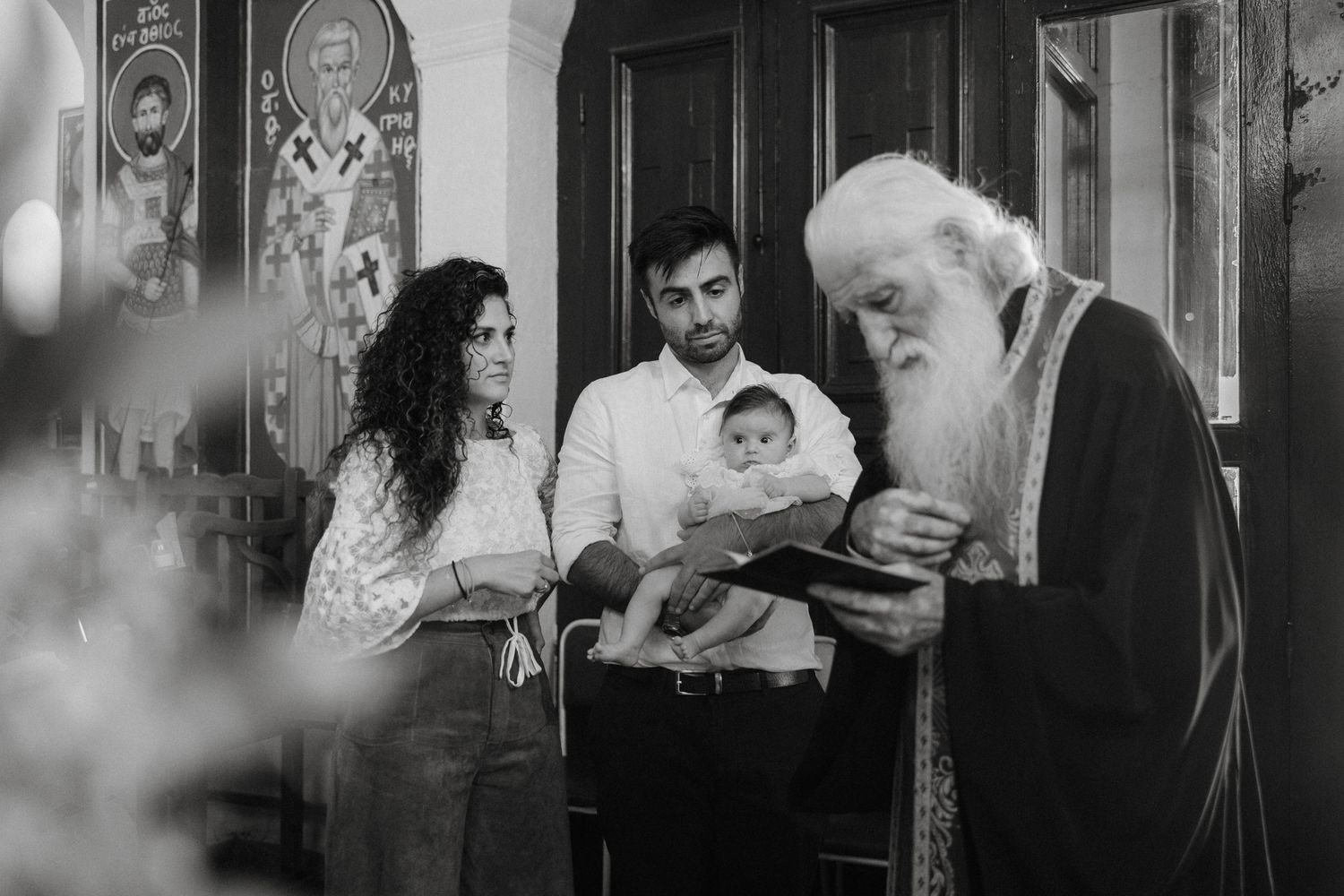 Black and white photo of a priest reading from a book during an Orthodox Christian baptism ceremony with parents and baby.