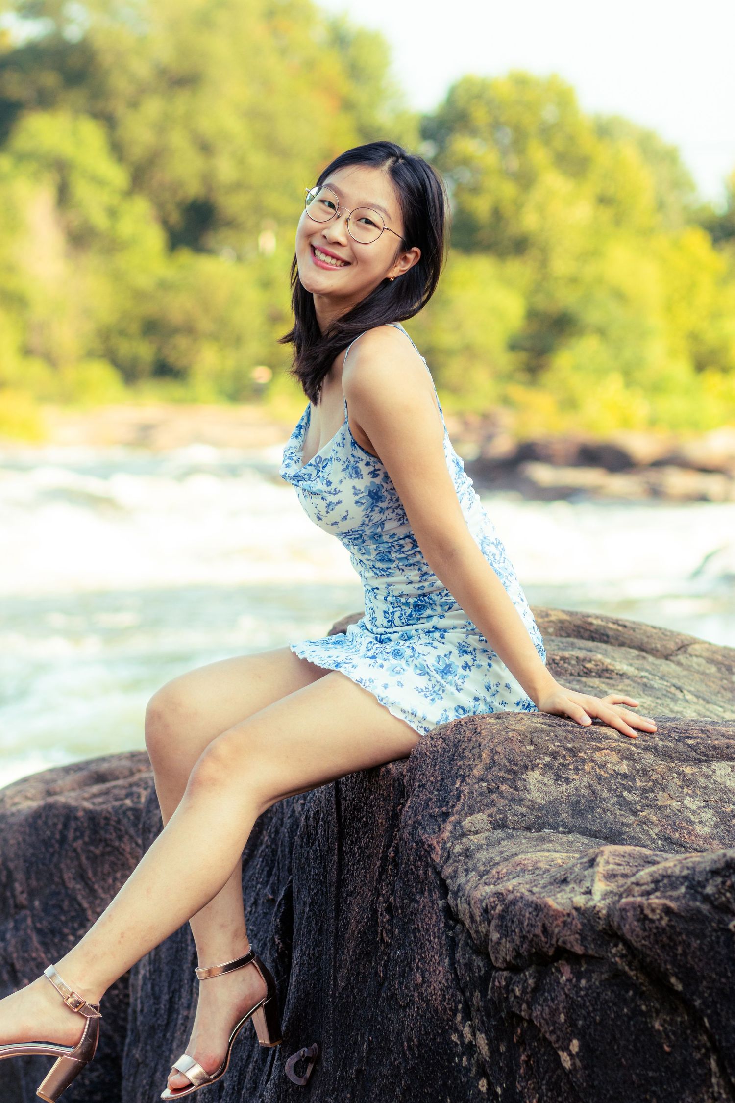 A person in a blue floral dress sits on a rocky outcrop near water on a sunny day.