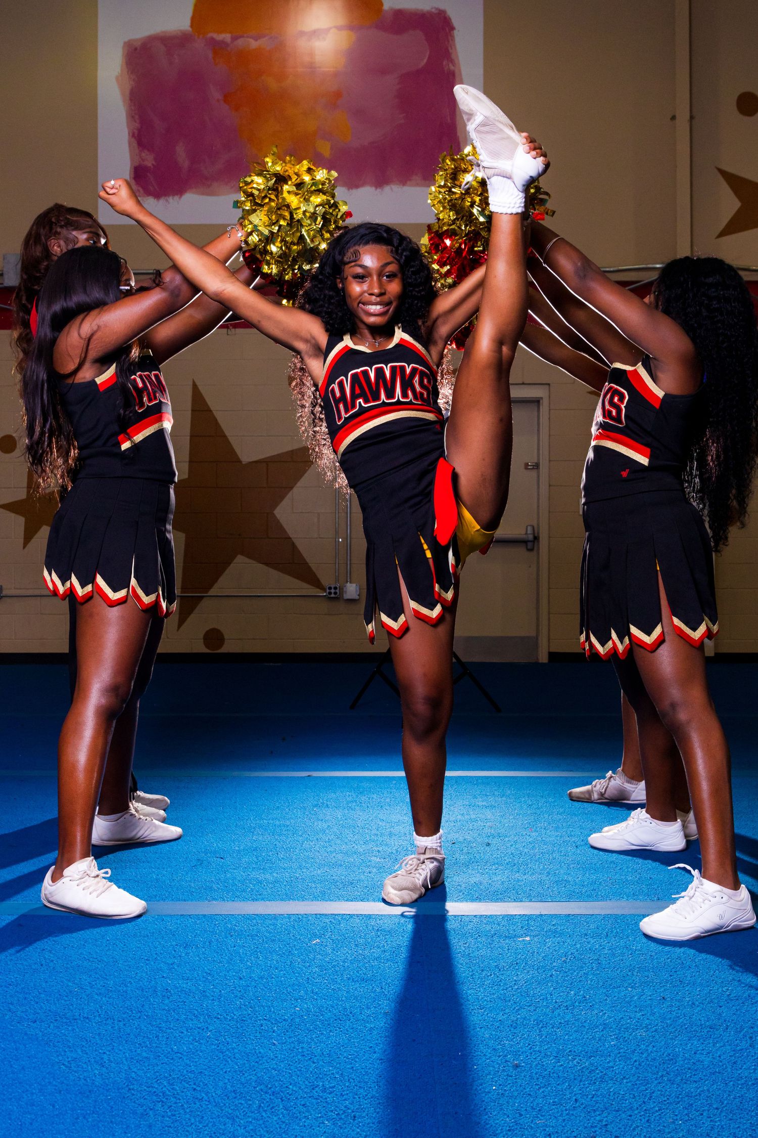 Cheerleaders in black and red uniforms performing high kicks and stunts on a blue floor mat during a routine.