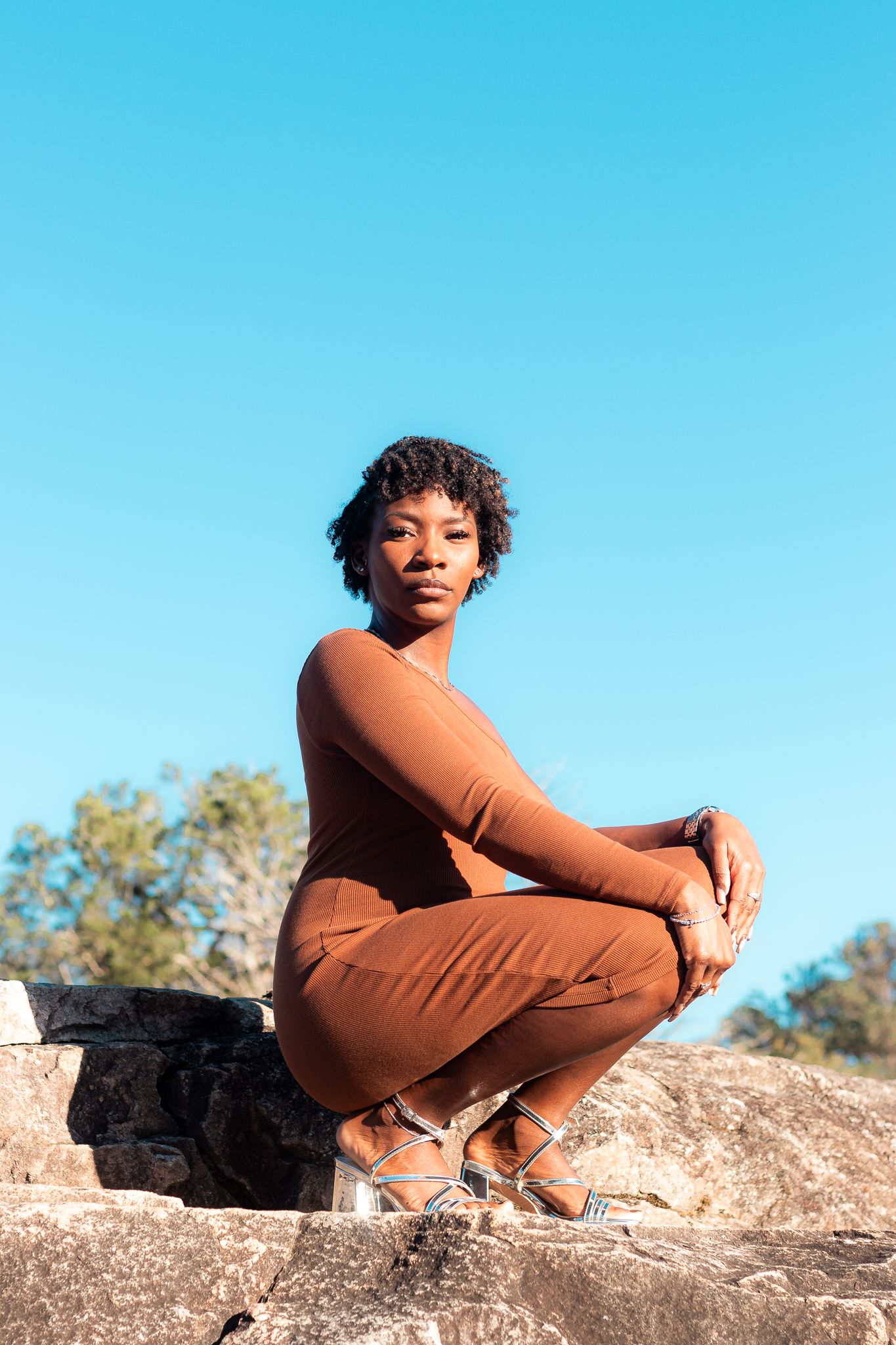 Model in earth tones crouches on natural rock formation with blue sky background.