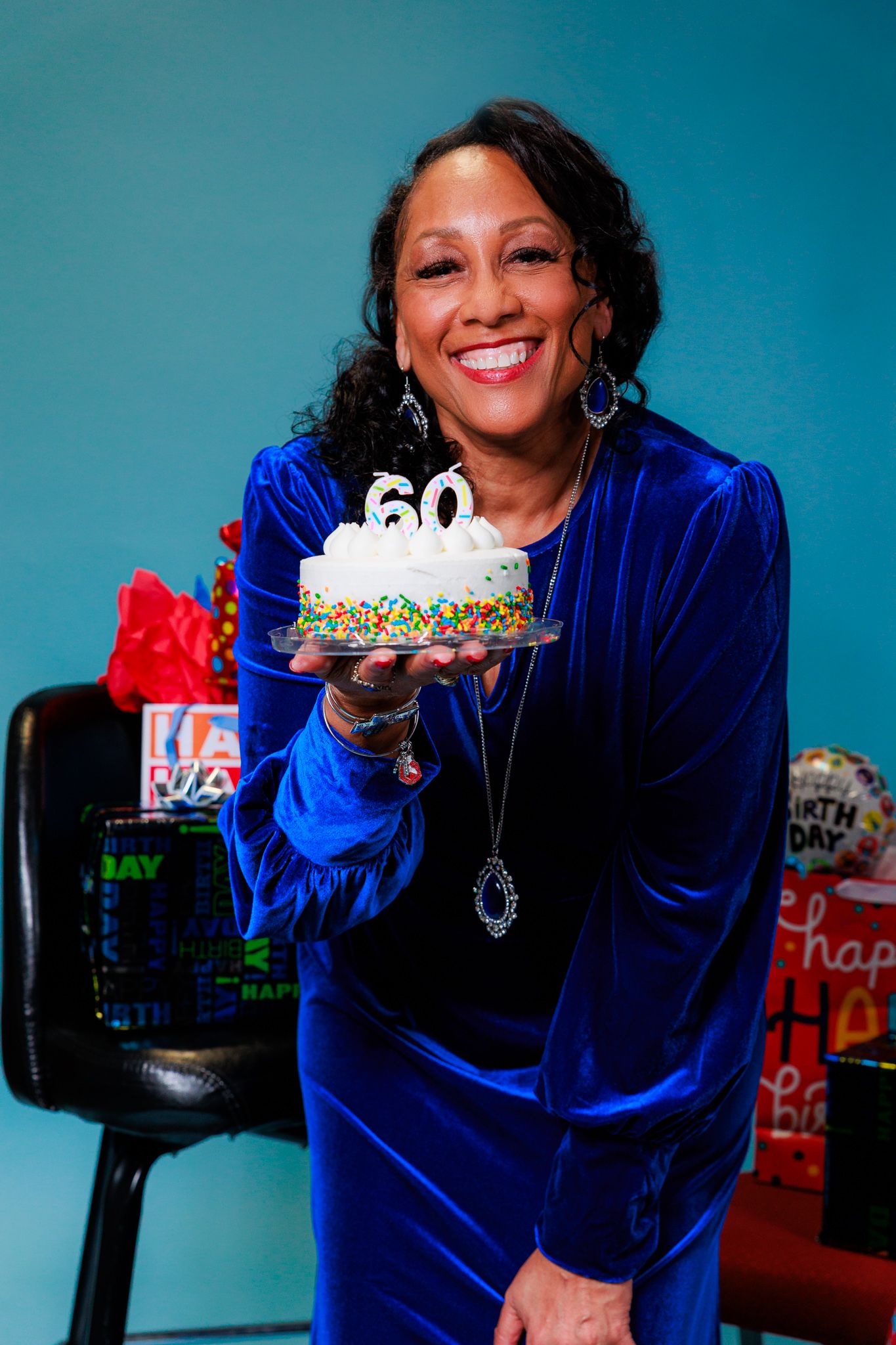 Someone in a royal blue velvet dress holds a birthday cake and gift bags against a turquoise background.
