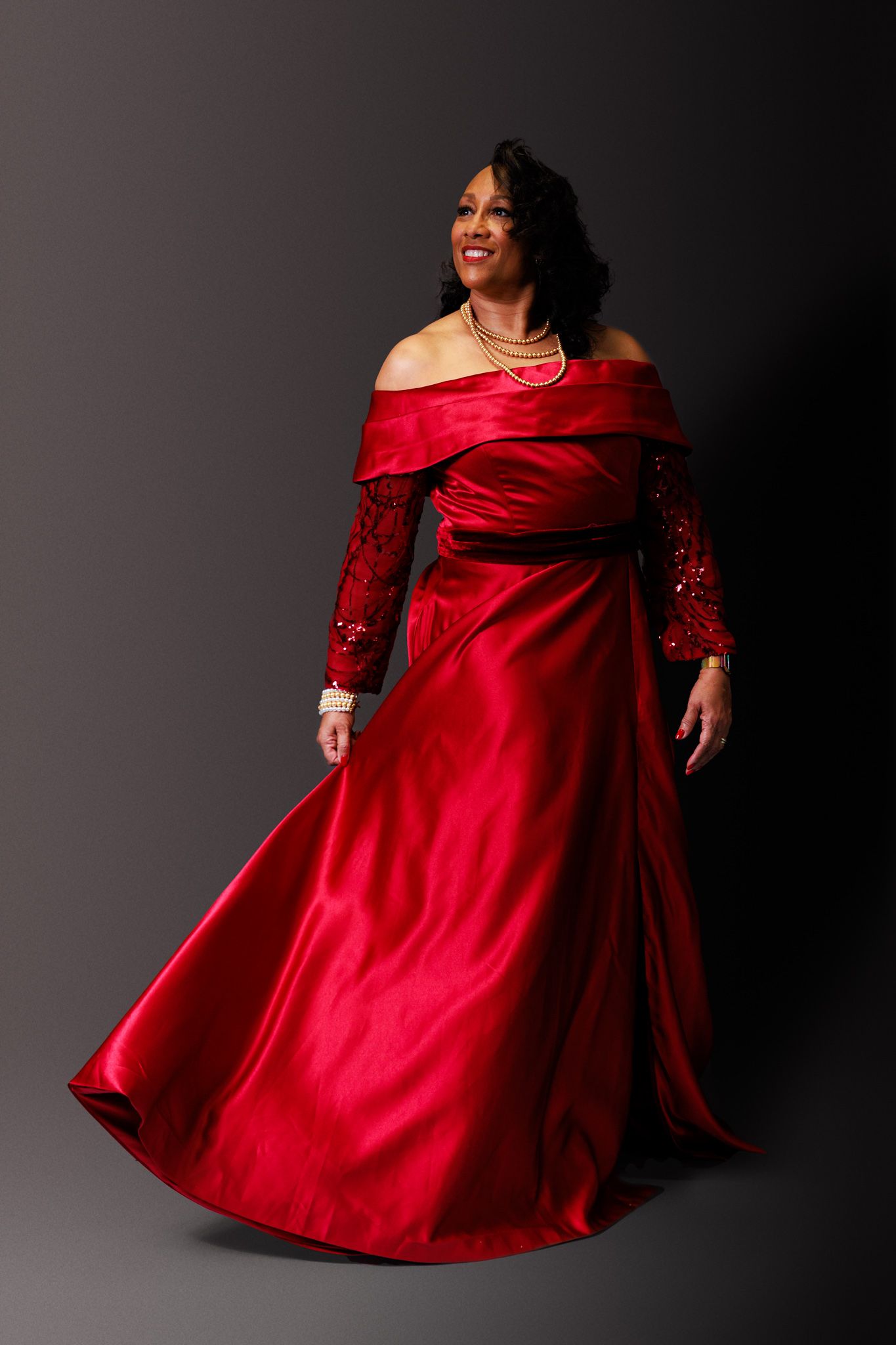 A model twirls in a flowing red formal gown with off-shoulder design against a dark studio background.