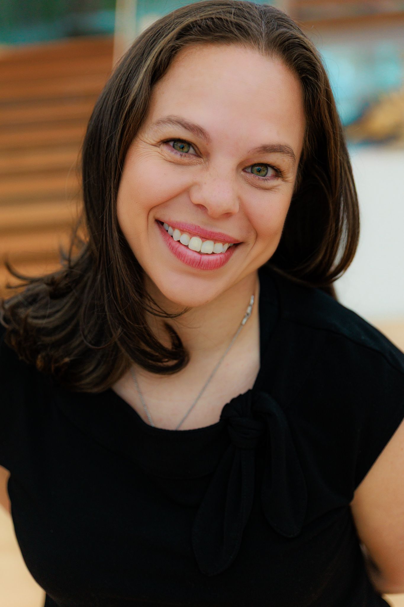 Smiling woman with dark hair wearing a black top and delicate necklace, posing for a professional headshot photo.