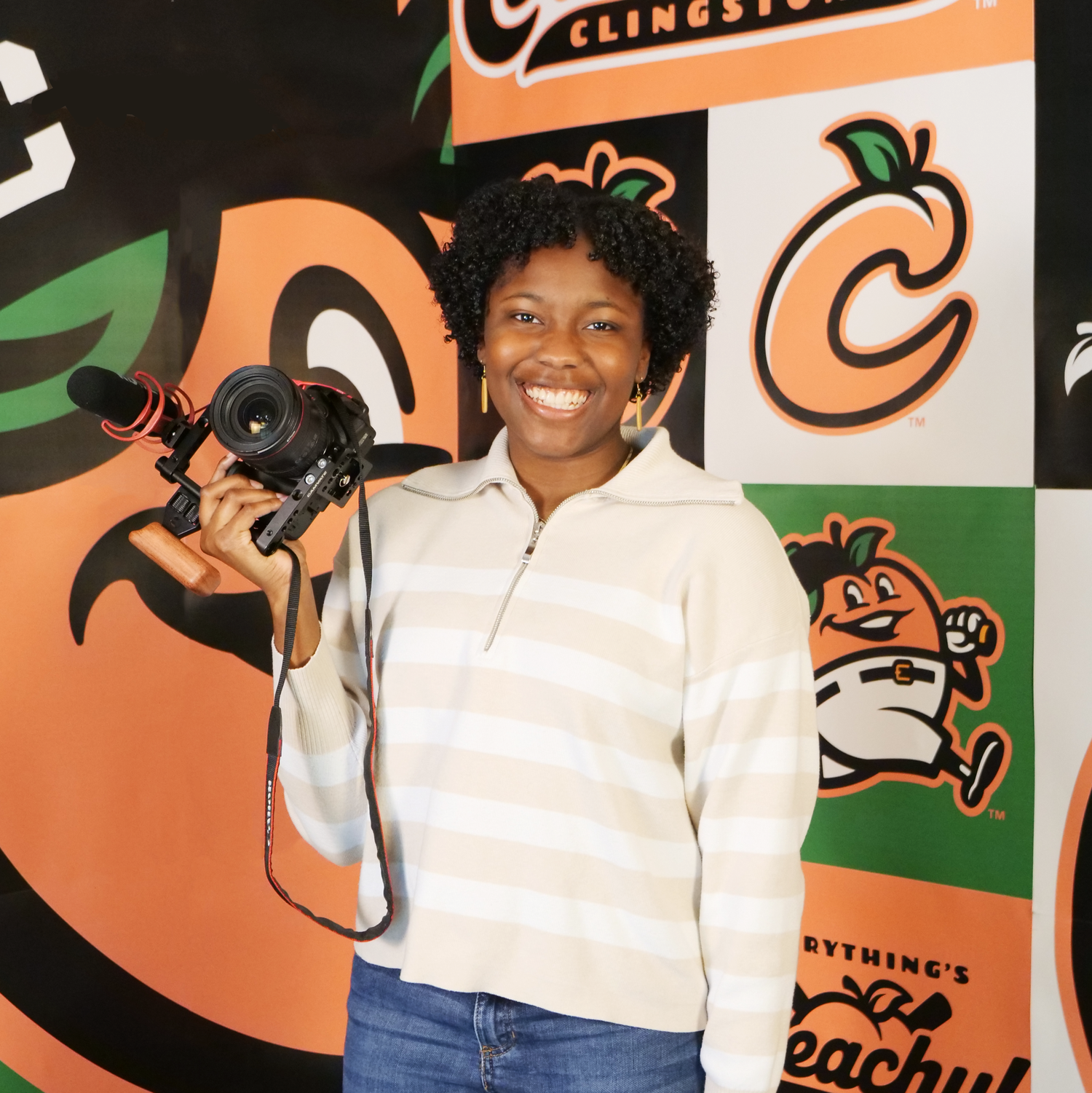 A person wearing a cream sweater holds a camera while smiling in front of a wall with orange and black cartoon graphics.