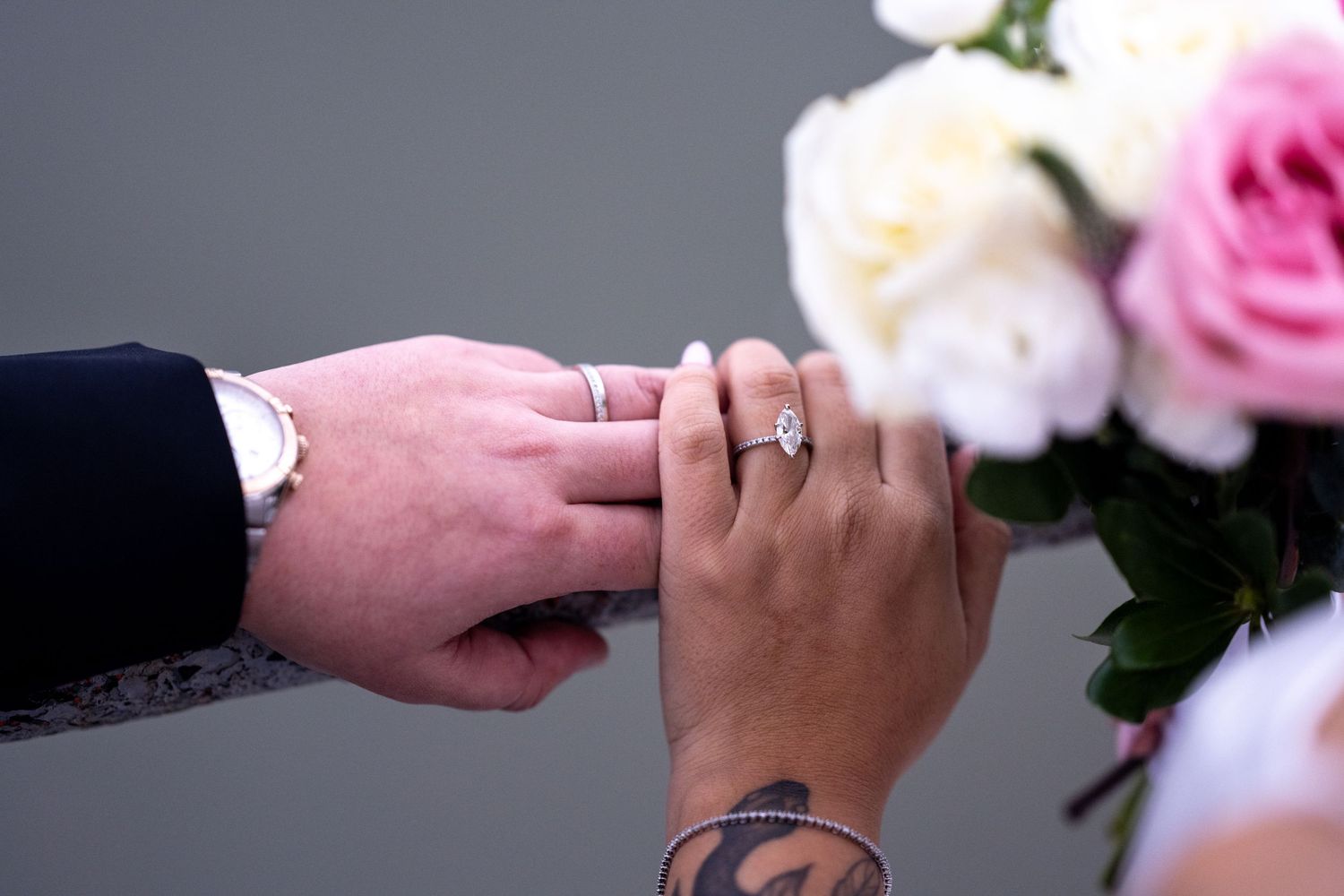 Groom placing a diamond engagement ring on brides tattooed hand with pink and white roses bouquet in background.