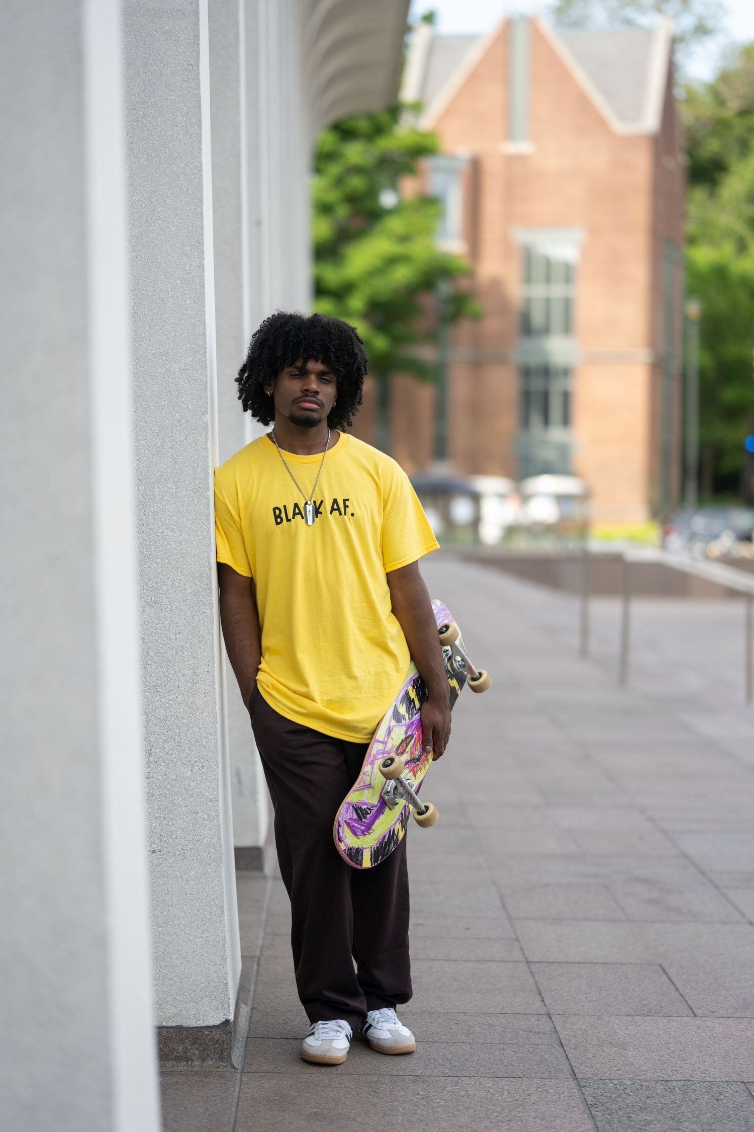 Skateboarder in yellow t-shirt and black pants holding skateboard while standing next to white wall on urban walkway.