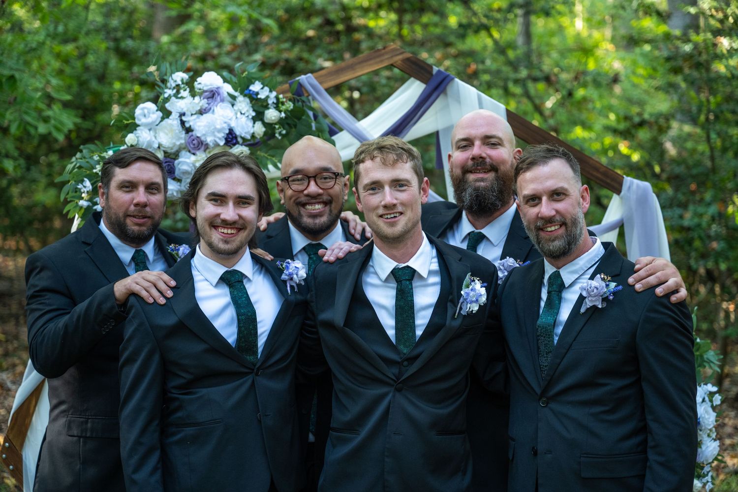 Groomsmen in black suits and ties pose together for formal wedding photos outdoors.
