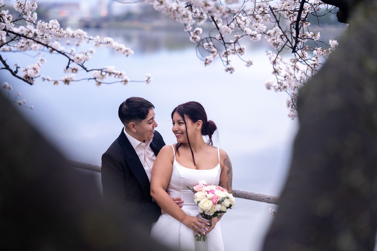 Couple in wedding attire embracing under cherry blossoms by a lake, bride holding a bouquet.