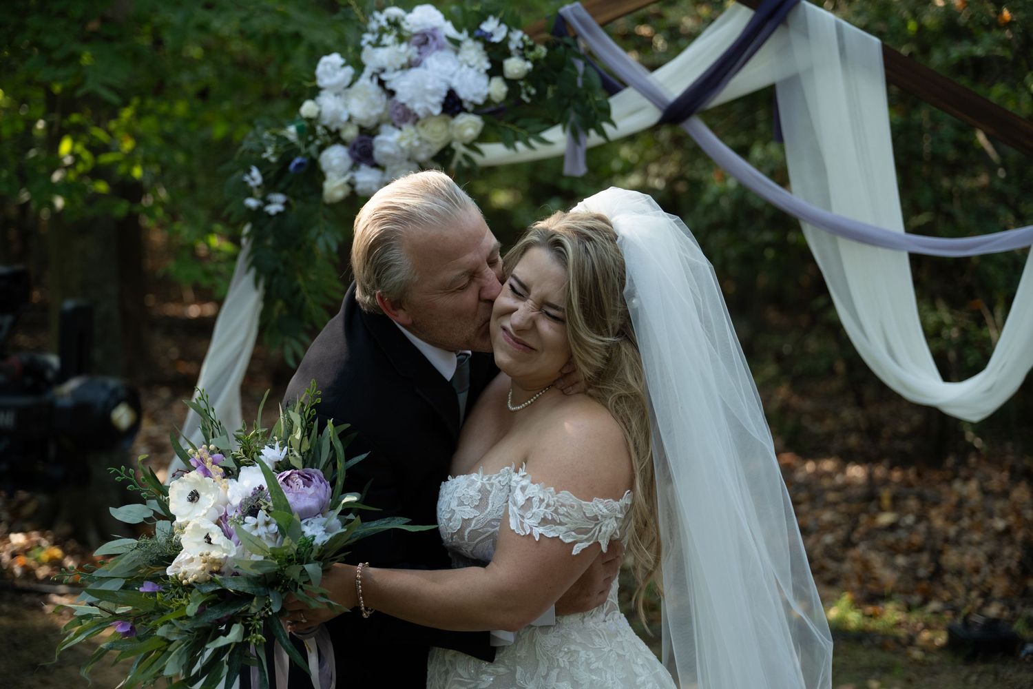 Wedding ceremony setup with white draped arch and floral arrangements in a wooded setting.