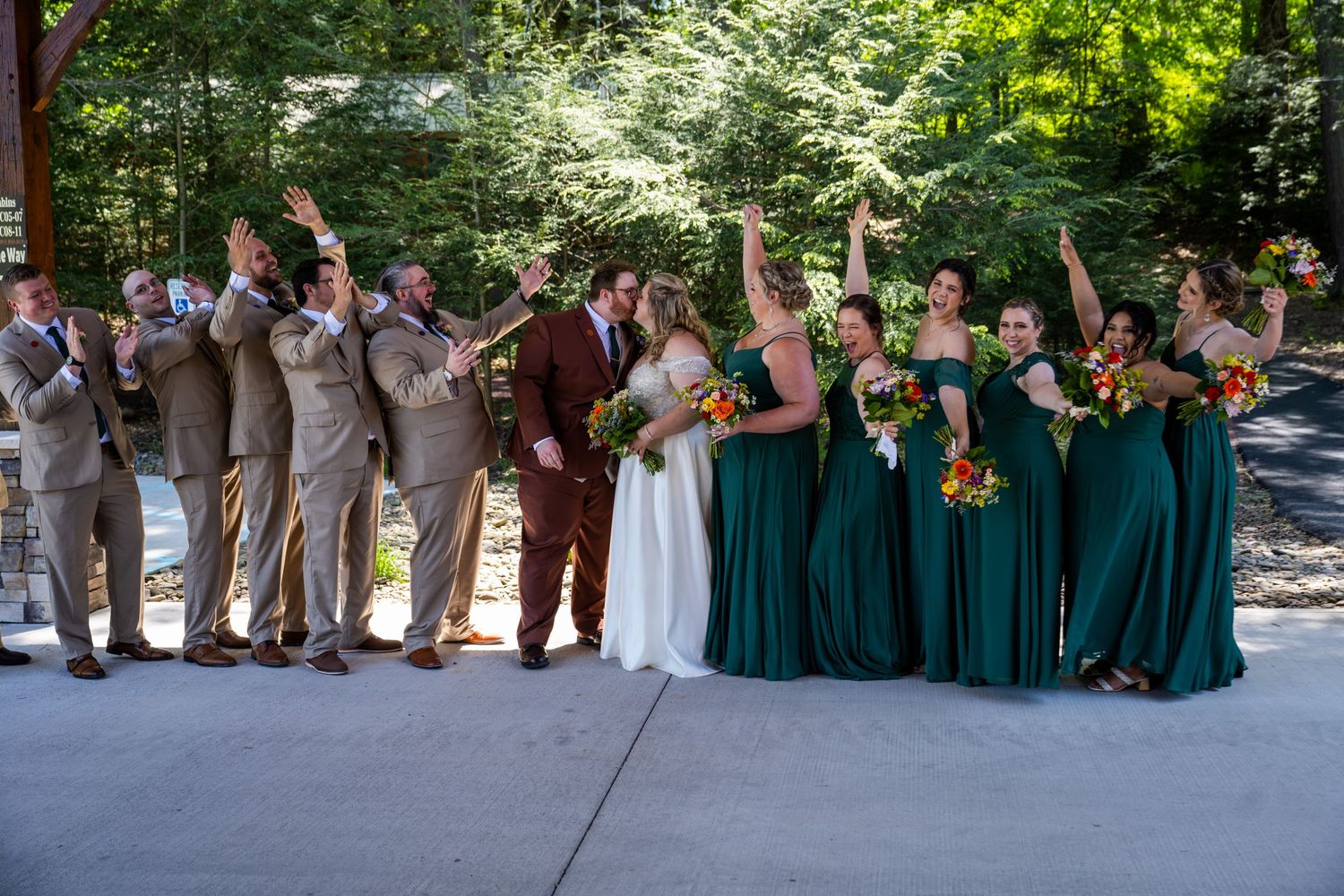 Wedding party in emerald green dresses and brown suits poses together on a paved outdoor path.