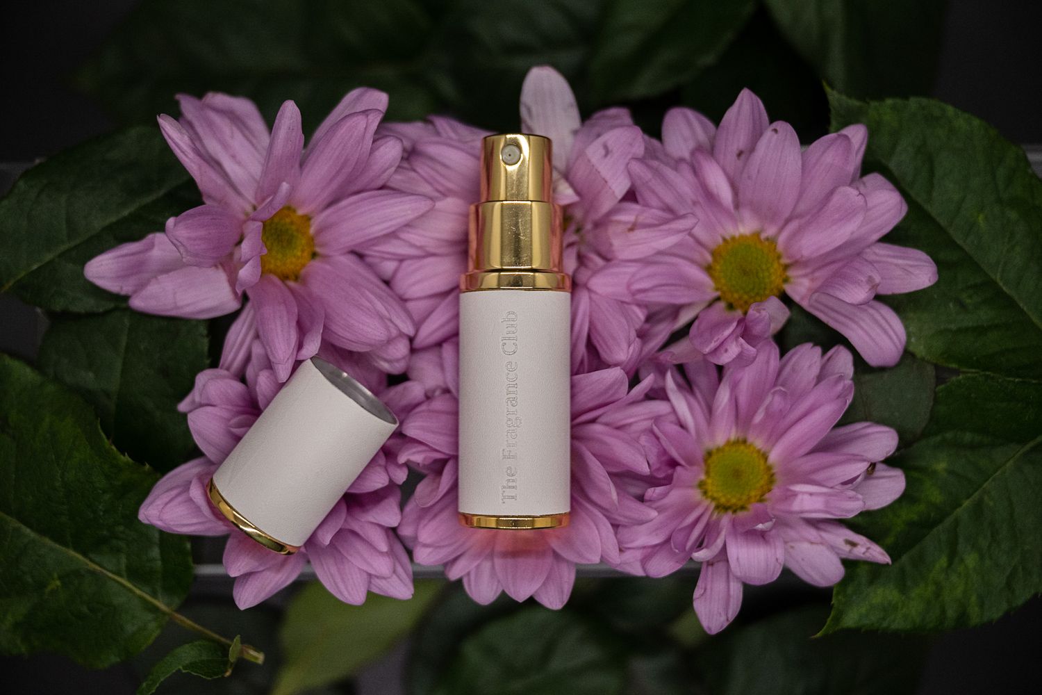 White cosmetic spray bottle displayed with pink daisy flowers and green foliage.