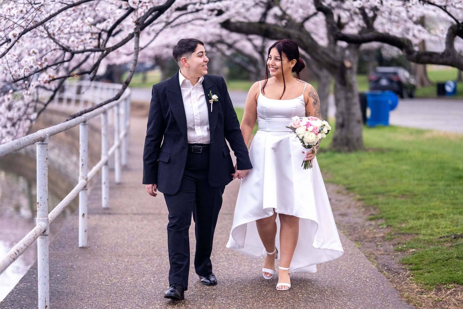 Couple in wedding attire walking hand-in-hand on a path lined with cherry blossom trees in full bloom.