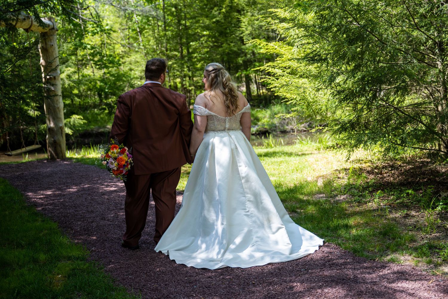 A bride in a white gown and partner in brown suit walk along a sunlit garden path surrounded by lush greenery.