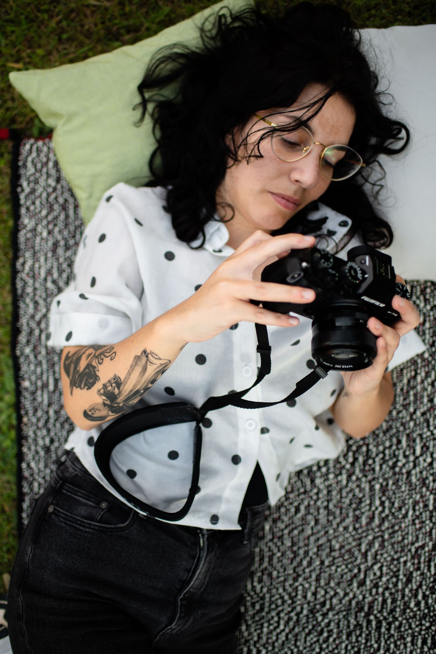 Photographer in polka dot blouse with floral arm tattoo lies on blanket while checking digital camera screen.