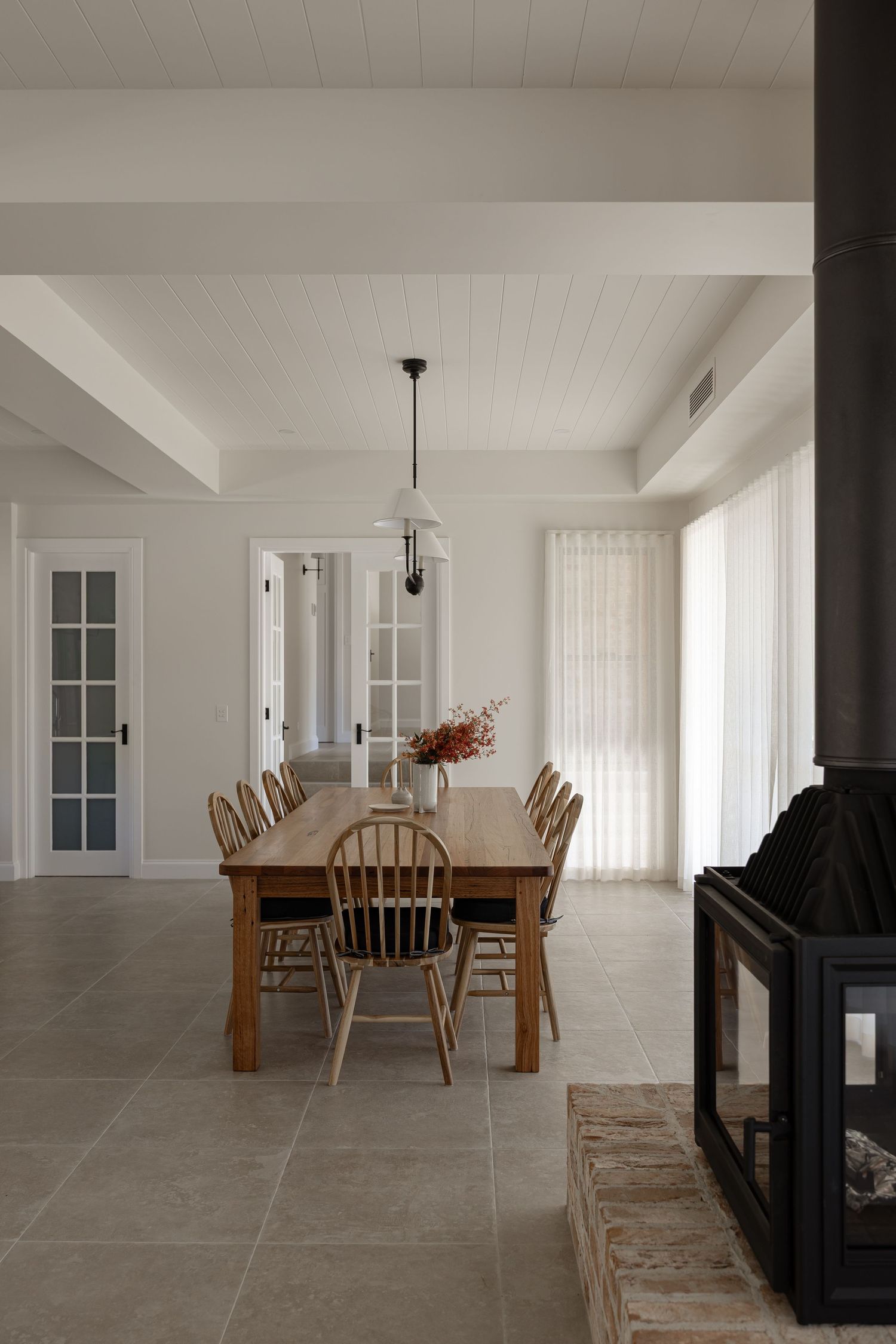 Modern farmhouse dining room with wooden table, Windsor chairs, white walls, and black fireplace in minimalist style.