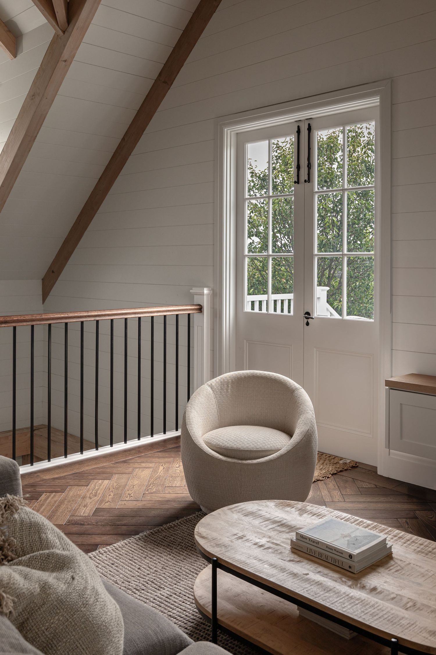 Modern attic reading nook with white walls, exposed wooden beams, and a cozy gray chair near windows overlooking trees.