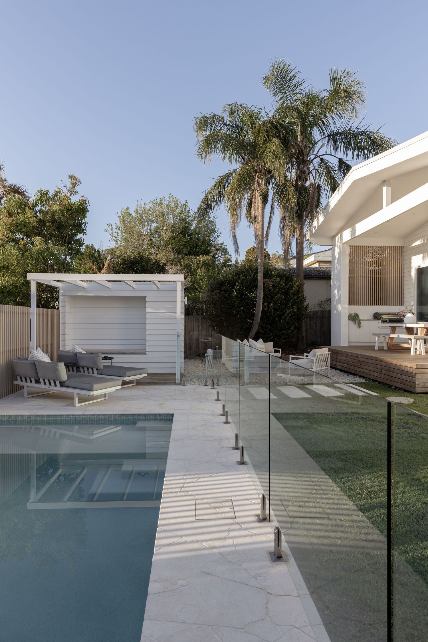 Modern poolside deck with glass railing, white concrete pavers, and palm trees in a luxury backyard setting.