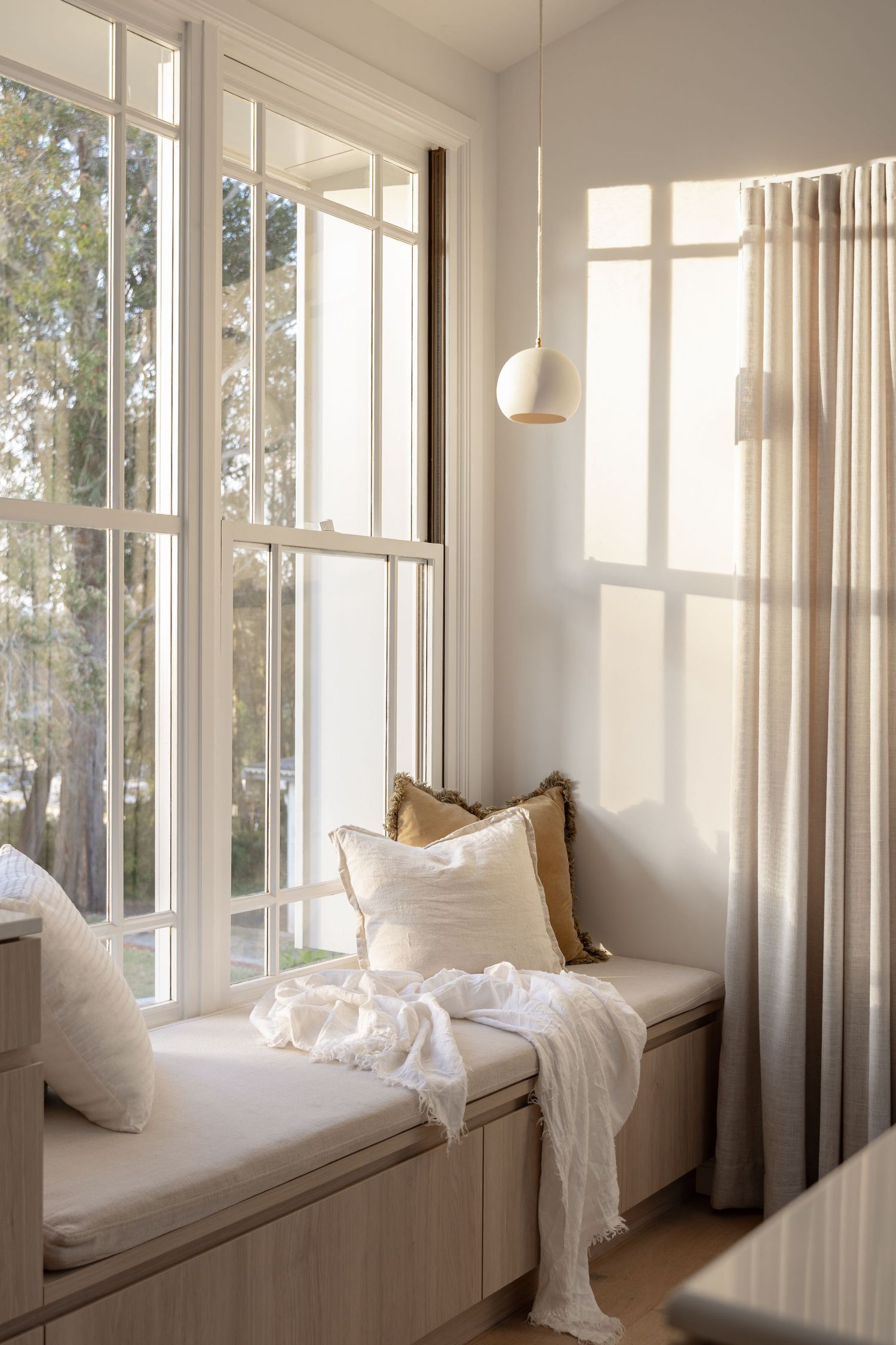 Cozy window seat nook with white cushions, throw pillows and sheer curtains bathed in natural sunlight.