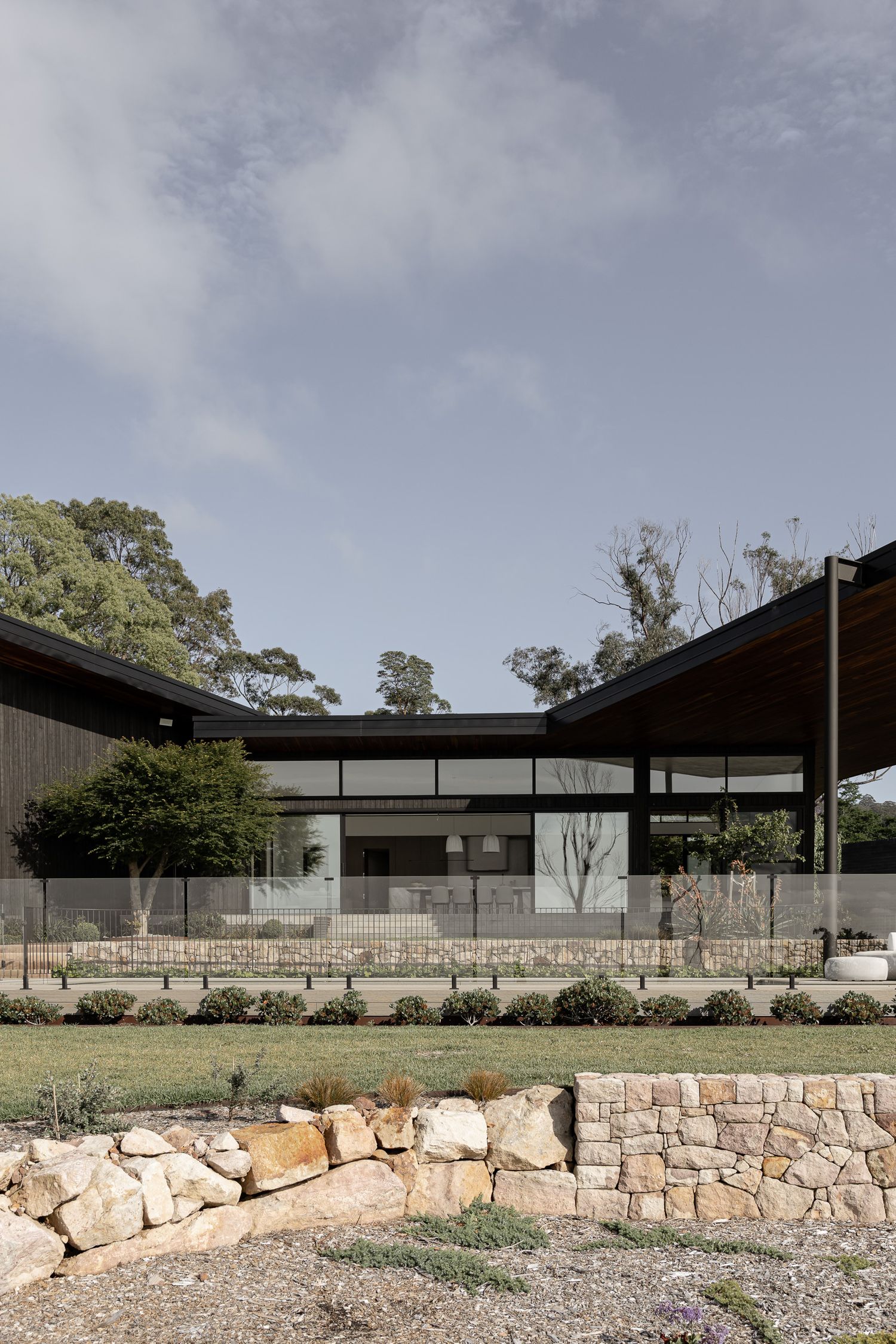 Modern Australian home with dark timber cladding, stone retaining walls, and native garden landscaping beneath a cloudy sky.