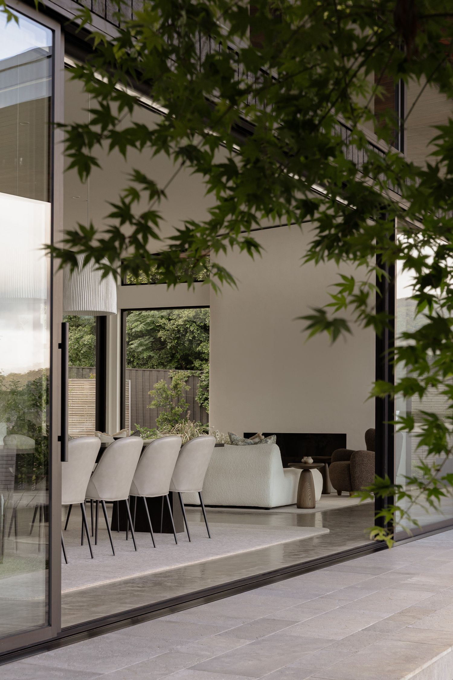 Minimalist dining area with cream chairs viewed through glass sliding doors framed by Japanese maple tree branches.