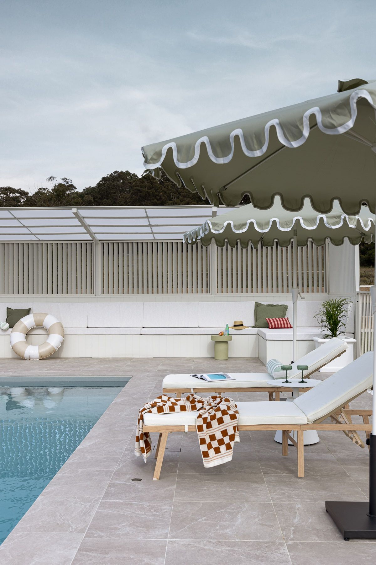 Poolside lounge area with white umbrellas, cushioned daybeds, and modern outdoor furniture arranged on a stone patio.