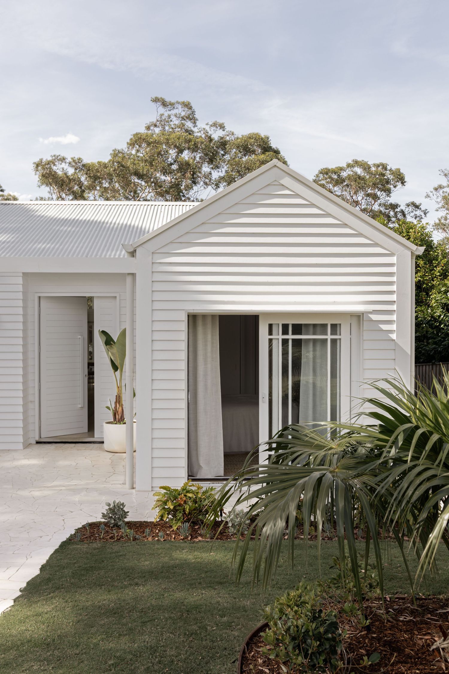 Modern white coastal home exterior with horizontal siding, gabled roof, and tropical landscaping in the front yard.
