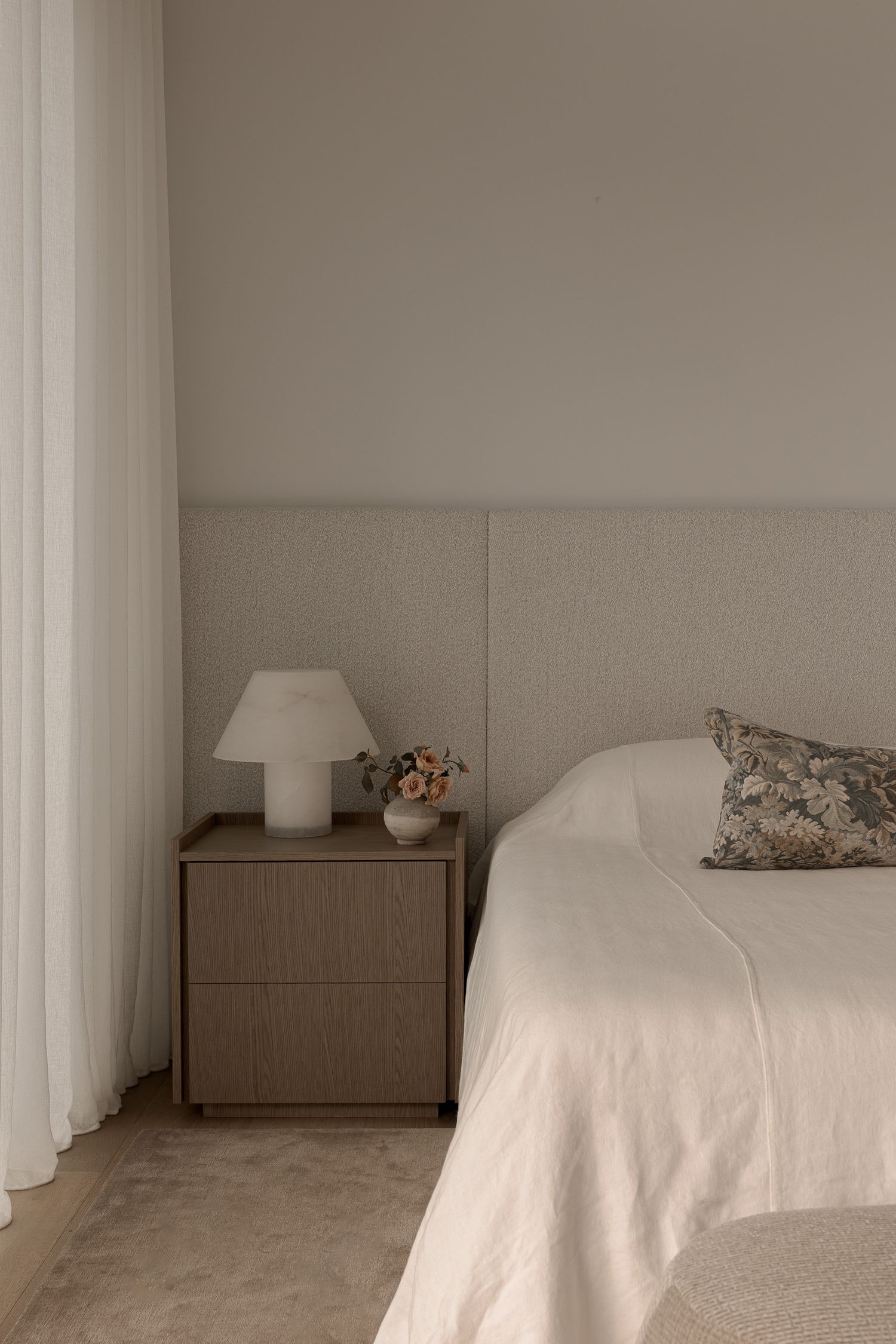 Minimalist bedroom with neutral tones, upholstered headboard, wooden nightstand, white lamp, and floral pillow accent.