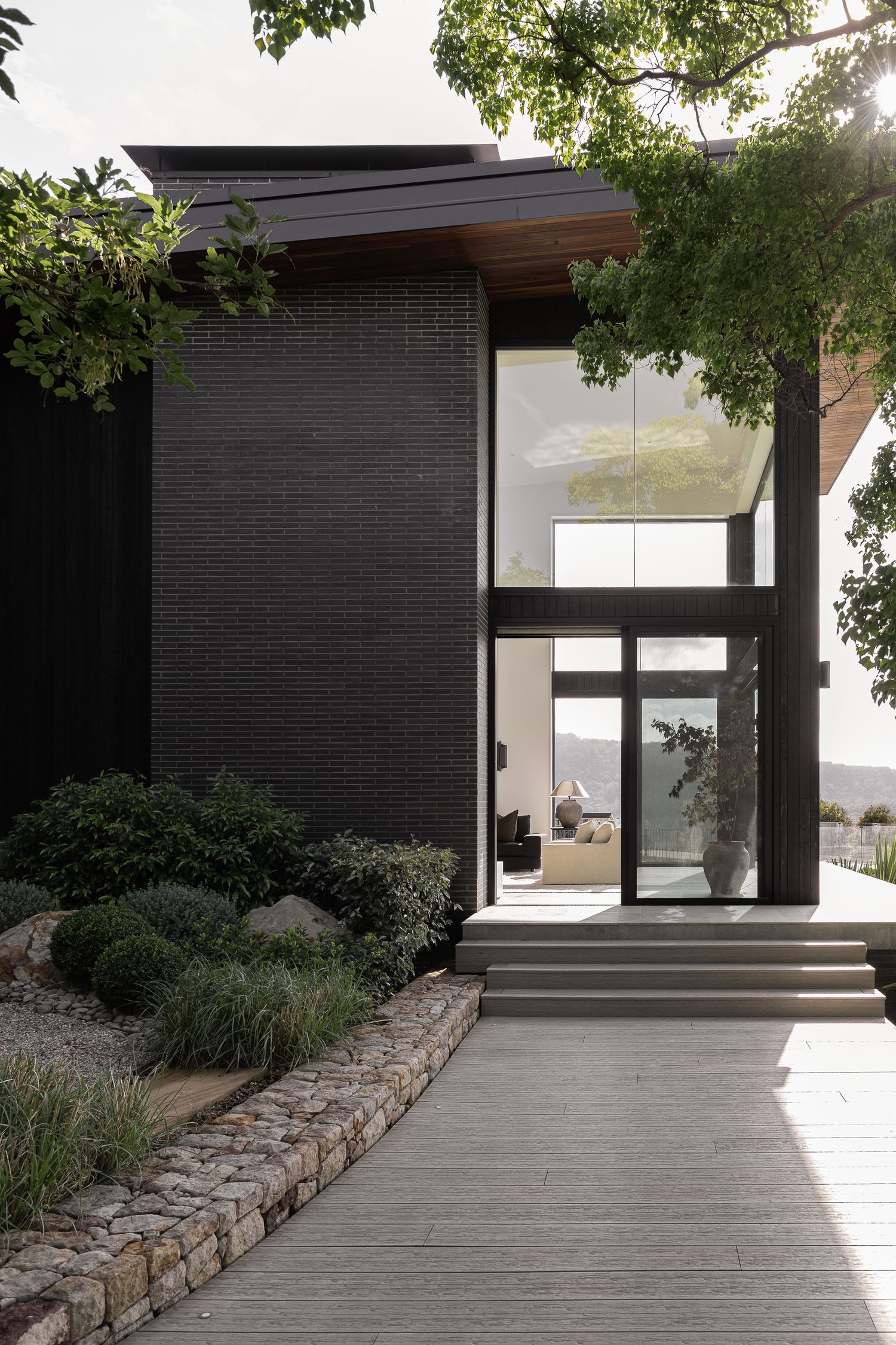 Modern black brick home entrance with glass doors, wood accents, stone garden border, and lush greenery in natural light.
