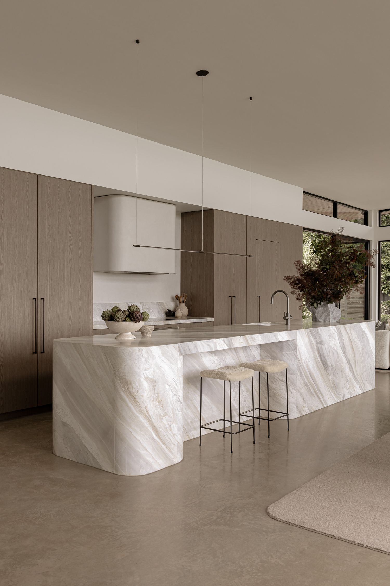 Minimalist kitchen with marble island, taupe cabinetry, concrete floors, and natural light from floor-to-ceiling windows.