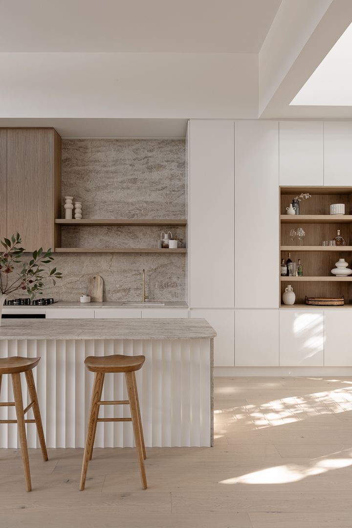 Contemporary white kitchen with fluted island detail and built-in shelving against textured stone wall.