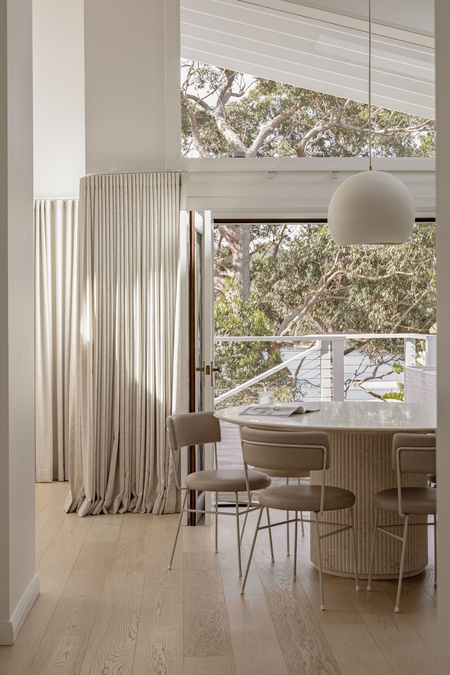 Modern dining area with beige curtains, curved chairs and dome pendant light features large windows overlooking trees.