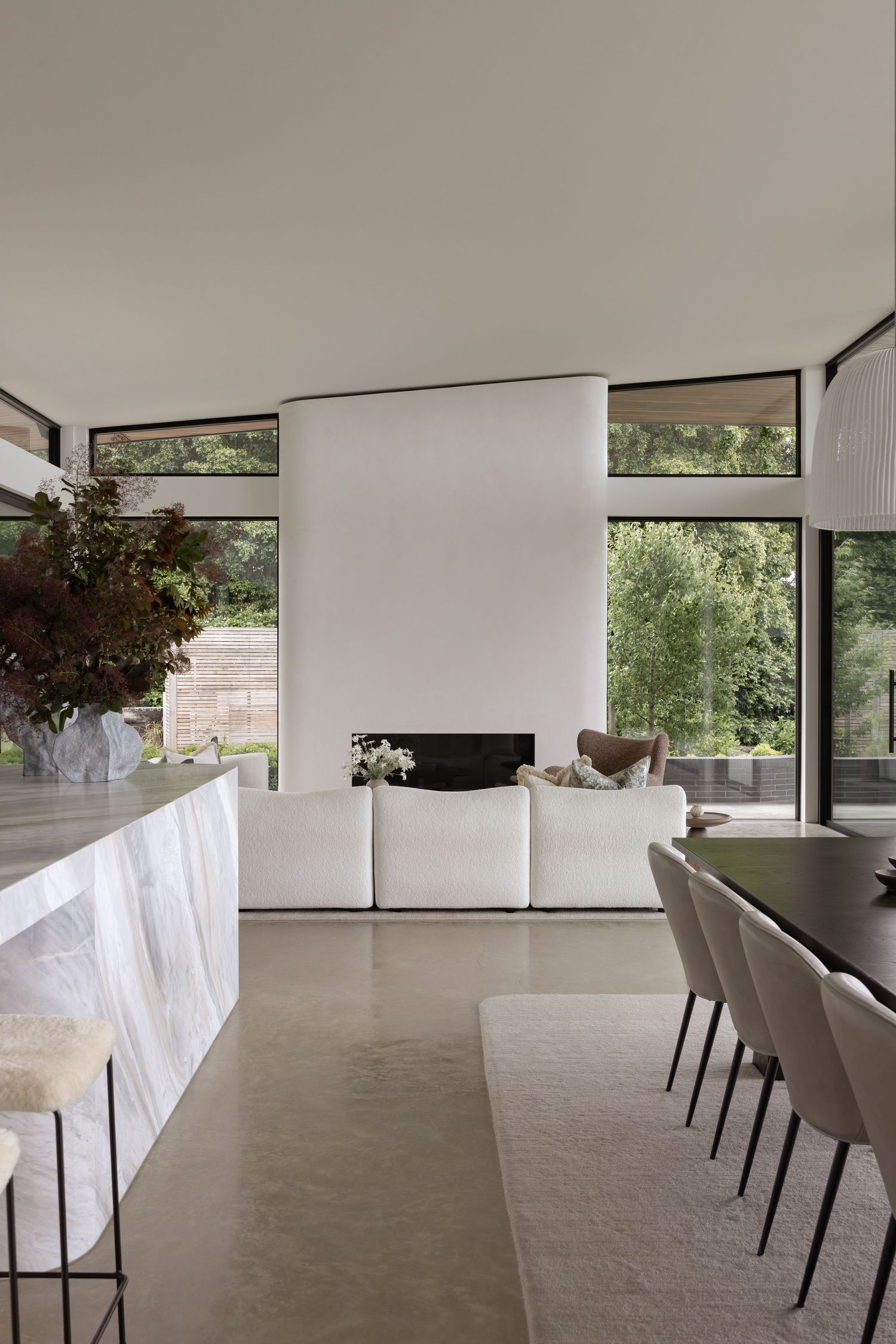 Modern minimalist dining room with white chairs and large windows overlooking greenery in a bright open space.