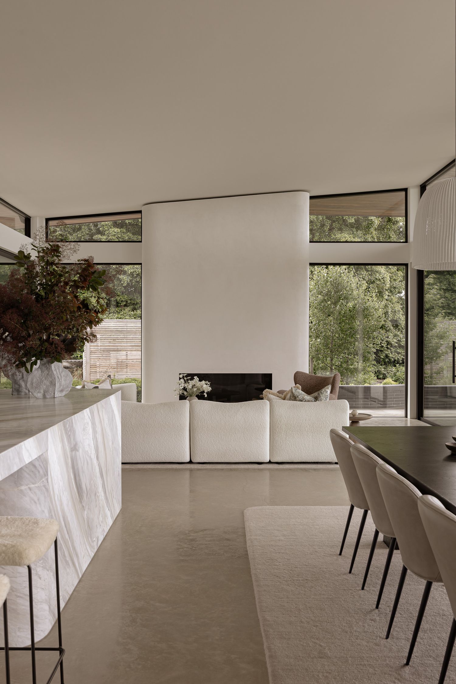 Modern open-plan living room with marble island, white sofa, fireplace, and floor-to-ceiling windows overlooking lush greenery.